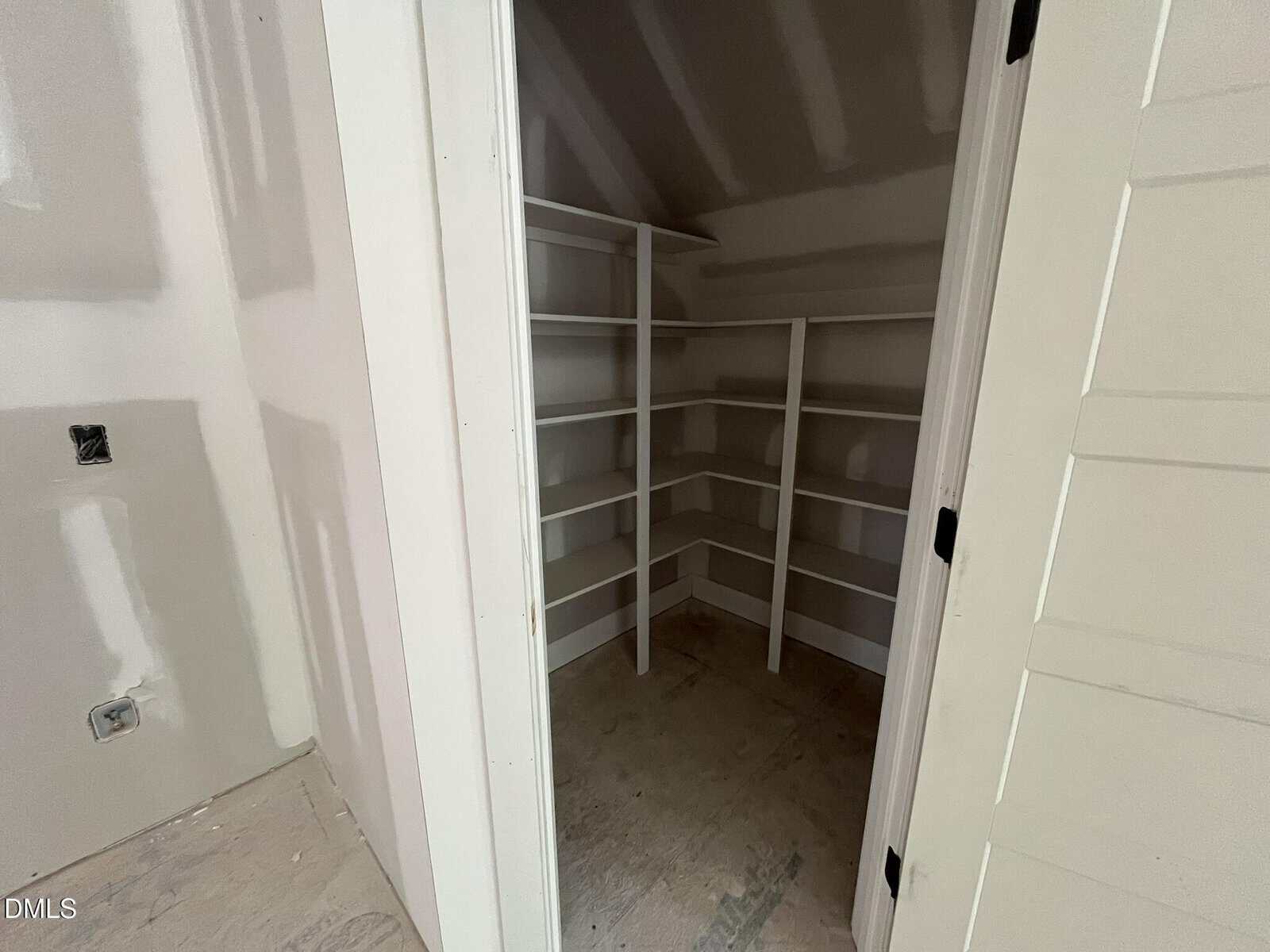 A walk-in closet with white shelves and unfinished drywall. The floor is bare, and the door is open, revealing angled walls and built-in shelving on three sides.