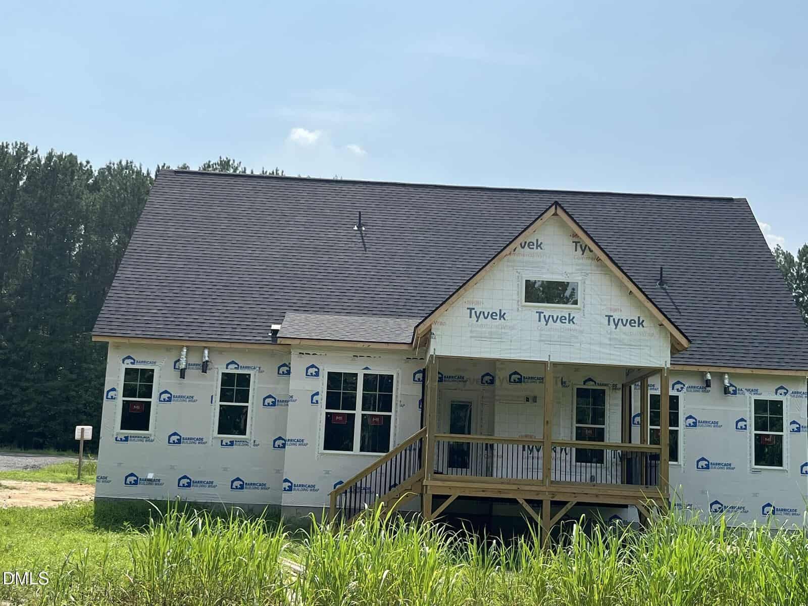 A partially constructed house with Tyvek wrap, black shingle roof, wooden porch, and surrounded by grass, stands in front of a forest under a clear blue sky.