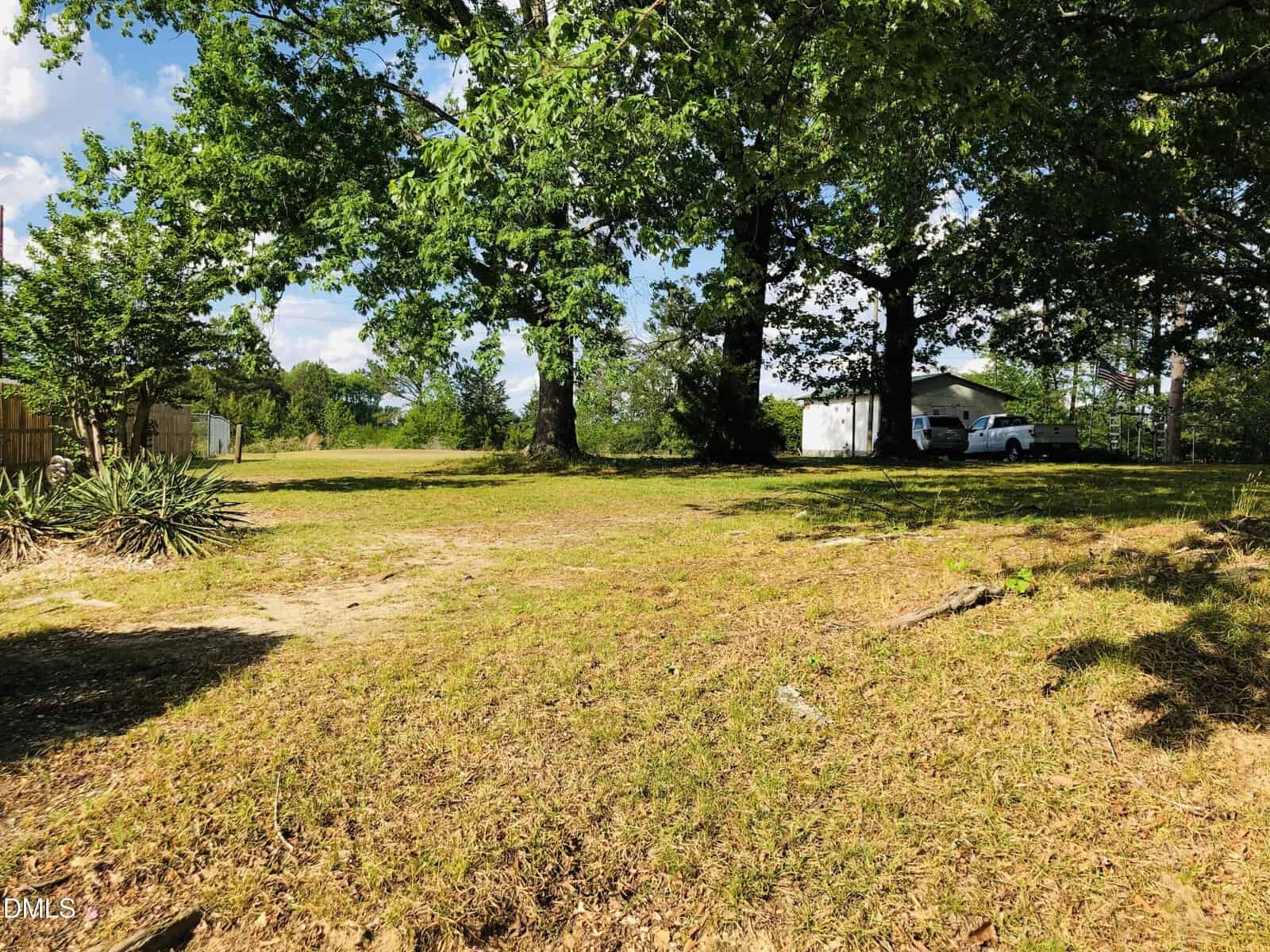 A grassy, sunlit yard with scattered trees and dirt patches along Tbd NC 56 in Franklinton, bordered by a wooden fence on the left. A white house, cars, and utility sheds are partially visible under a blue sky.