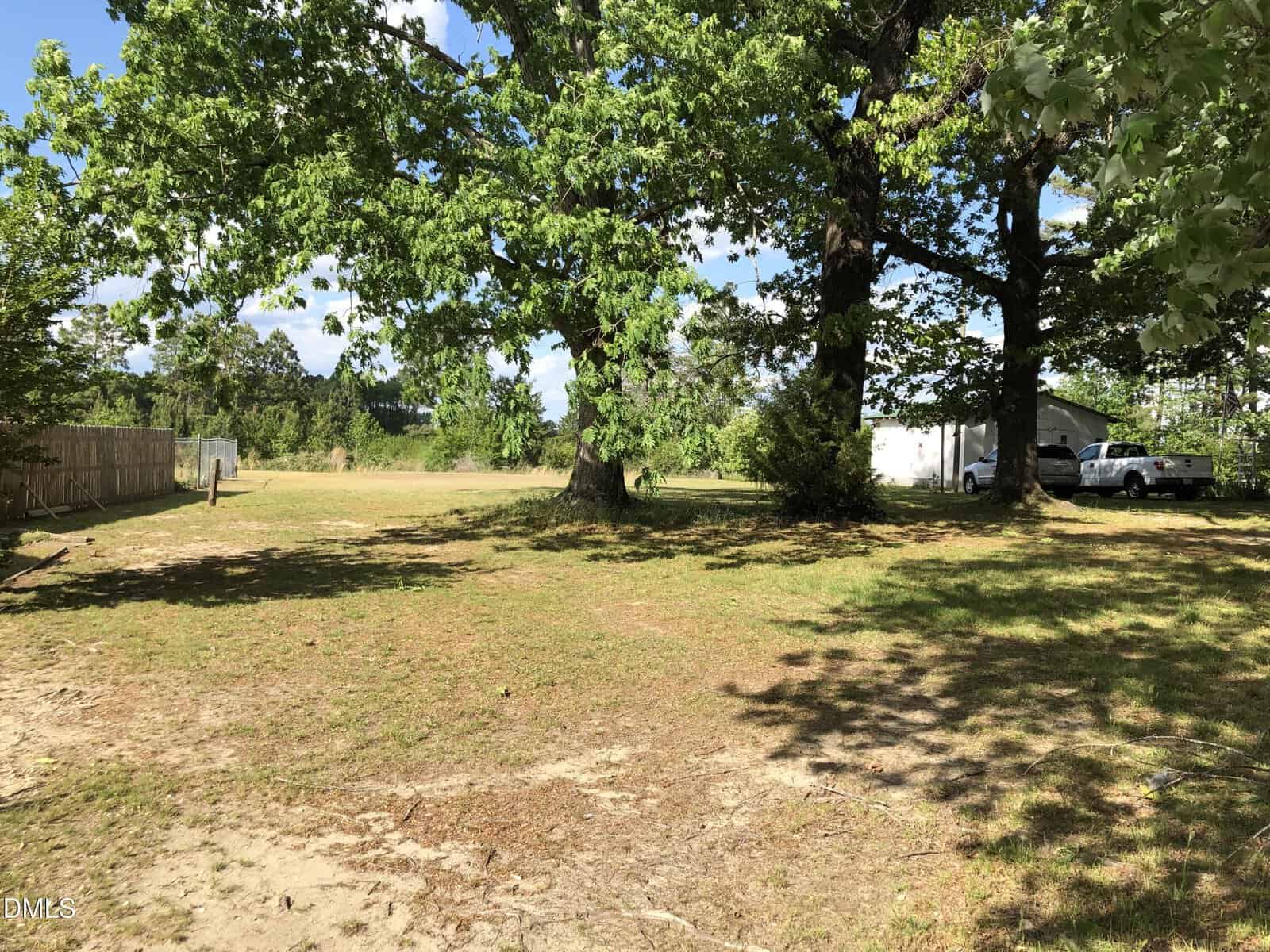 A sunny yard with patches of grass and dirt, scattered large trees providing shade, a wooden fence on the left, and a white building with vehicles parked nearby in the background.