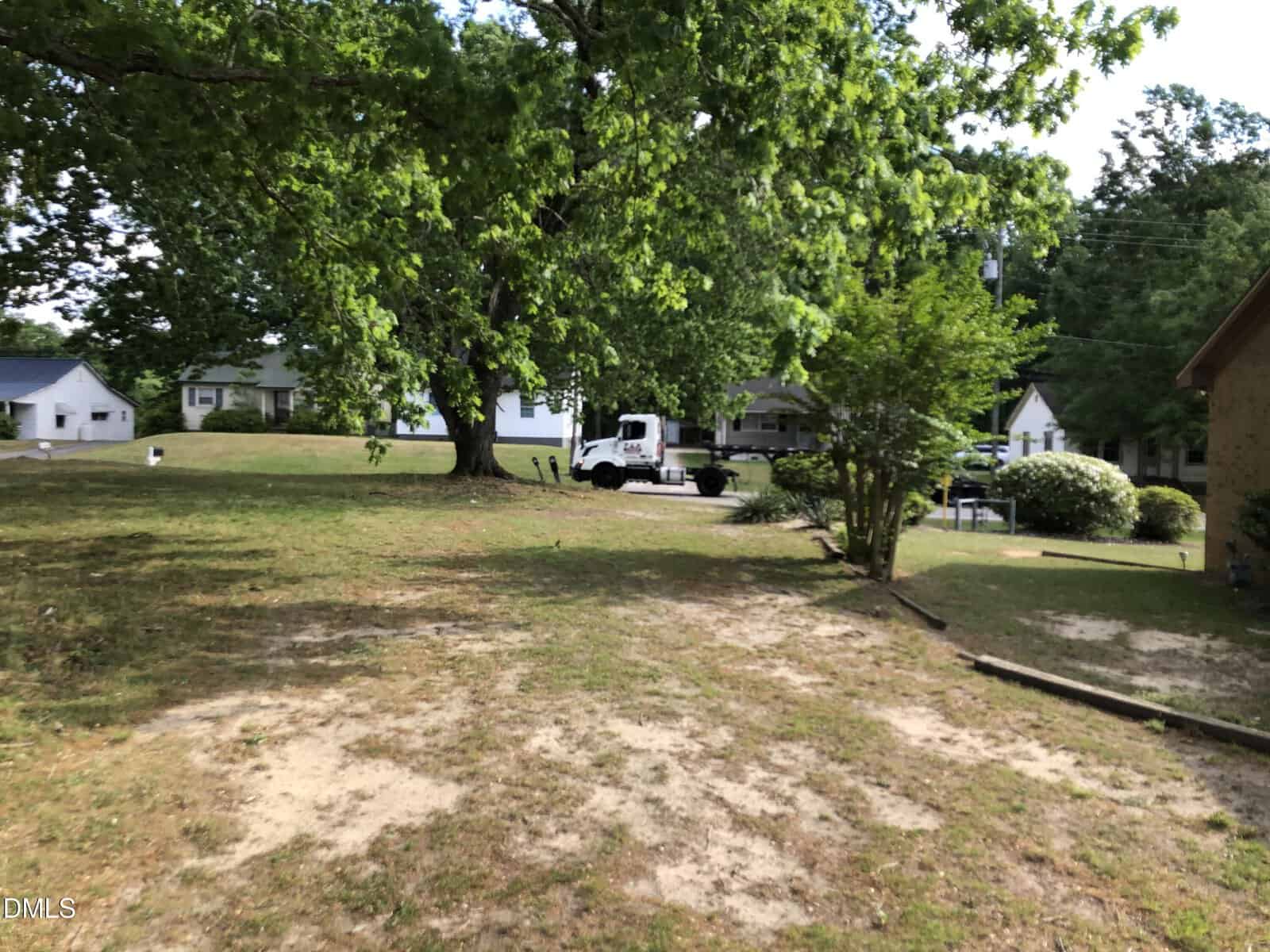 A grassy yard with patches of bare soil, large leafy trees, and a white truck parked near the center. Several houses are visible in the background, surrounded by greenery.