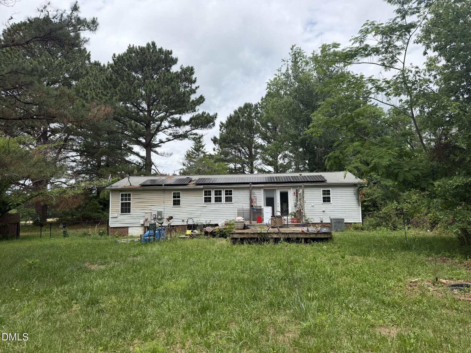 A single-story white house with a large, overgrown yard, surrounded by tall trees. The house has a deck with various items scattered on it and appears weathered and in need of maintenance.