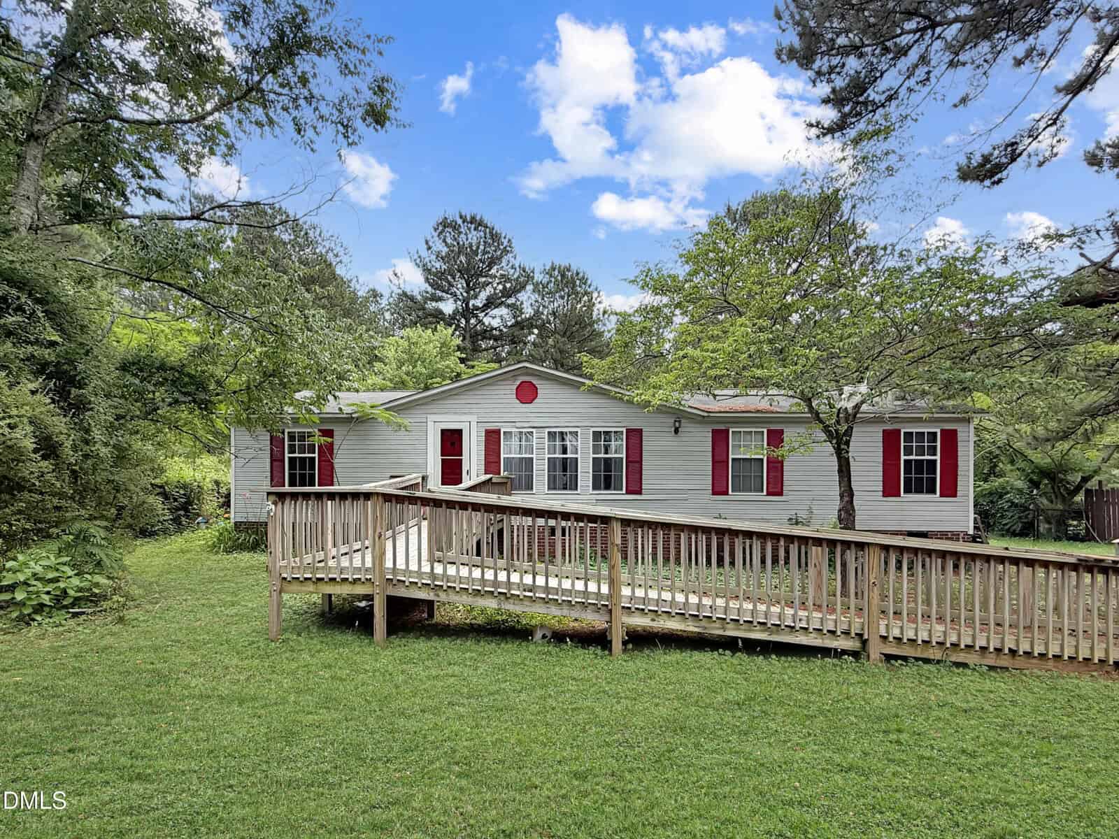 A single-story house on 4298 Tommie Sneed Road in Oxford features gray siding, red shutters, and a wooden ramp leading to the front door, all set amid green grass, trees, and a blue sky with scattered clouds.