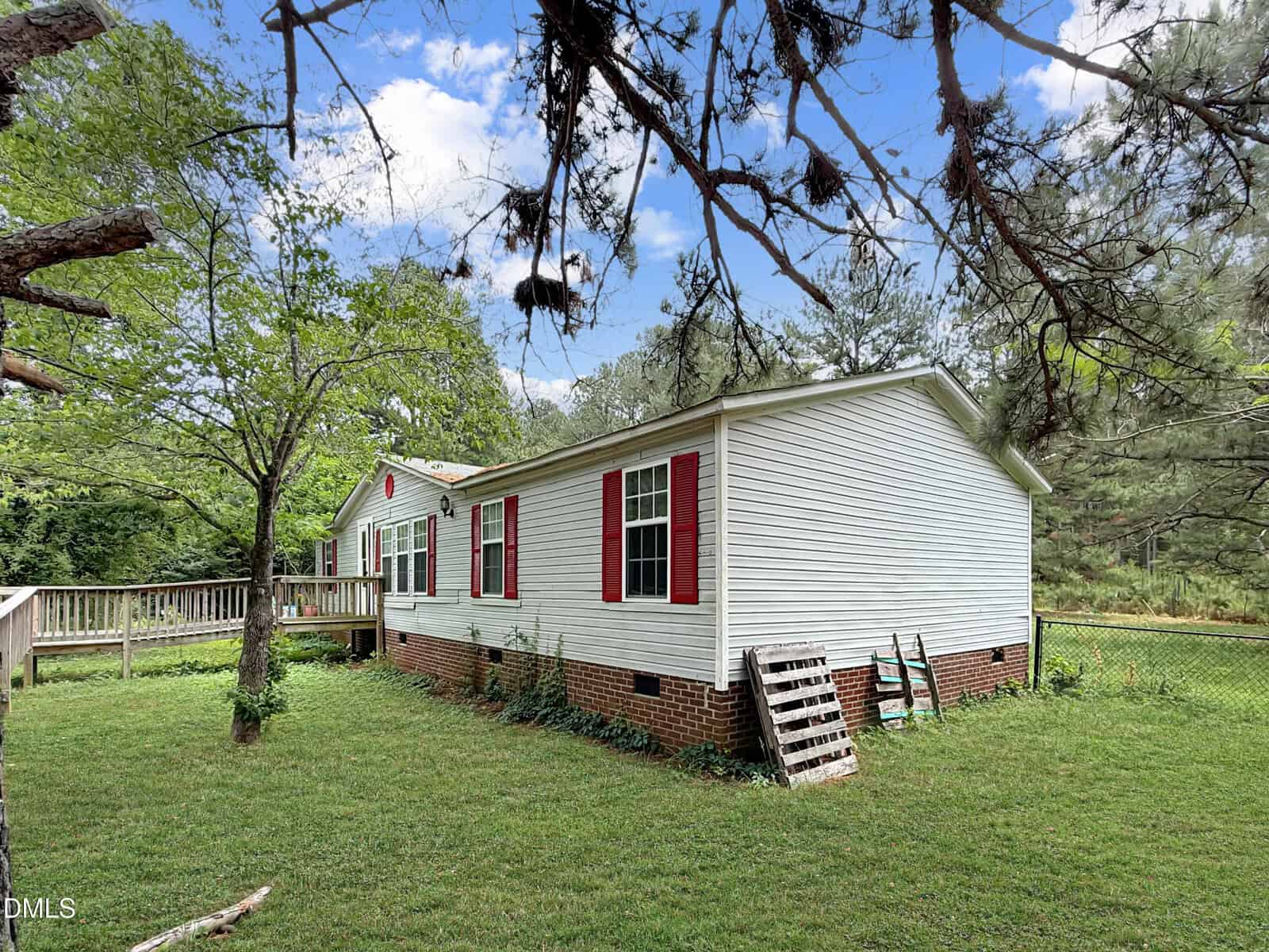 A white manufactured home with red shutters and a brick foundation sits on a grassy lawn, surrounded by trees and a chain-link fence under a partly cloudy sky. A wooden ramp and pallet lean against the house.