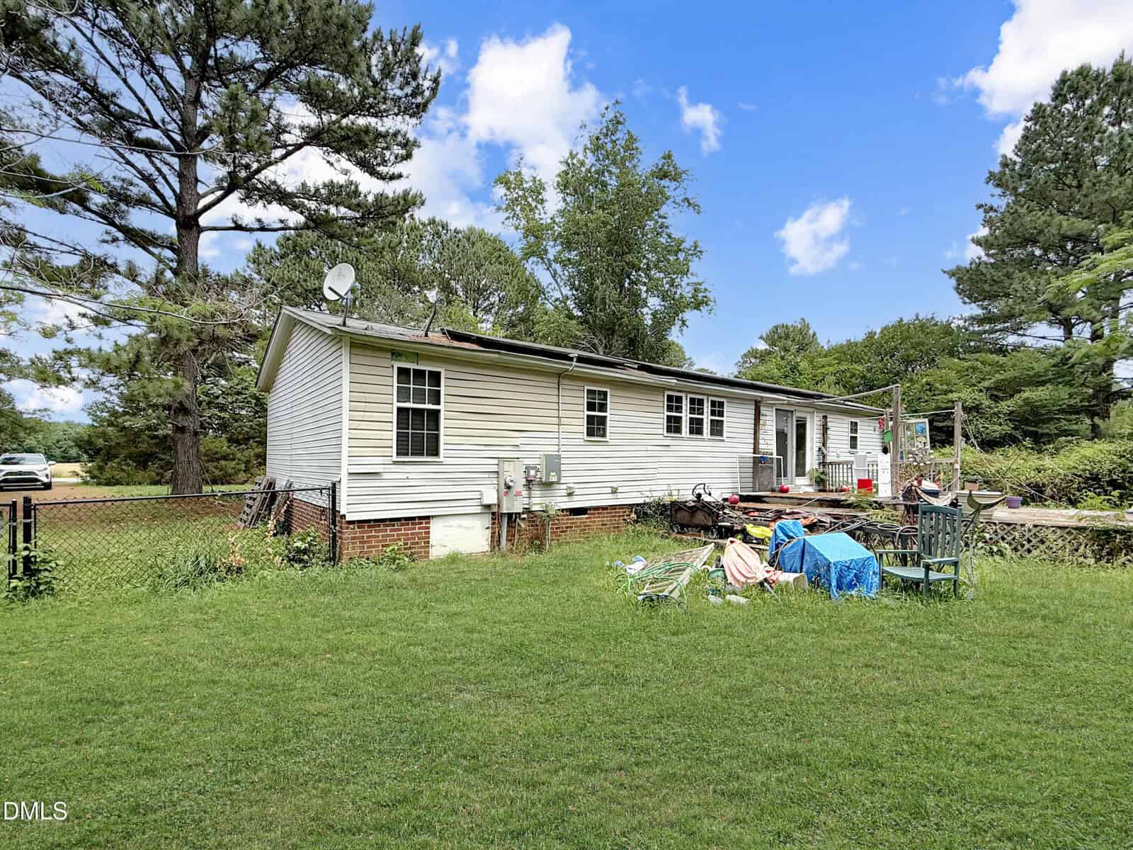 A single-story white house with a fenced yard, surrounded by tall trees. Outdoor furniture and various items are scattered on the grass, and the sky is blue with scattered clouds.