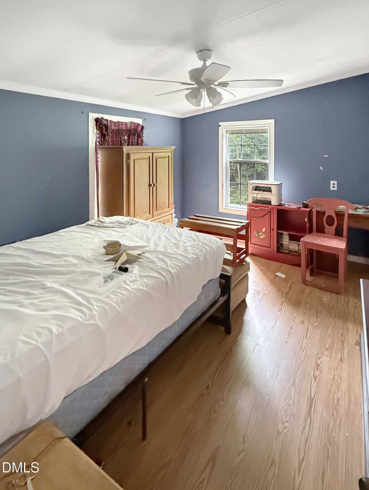 Bedroom with blue walls, a bed with white sheets, wooden furniture including an armoire, a red desk with chairs, and a window letting in natural light. Ceiling fan and wood flooring are visible.