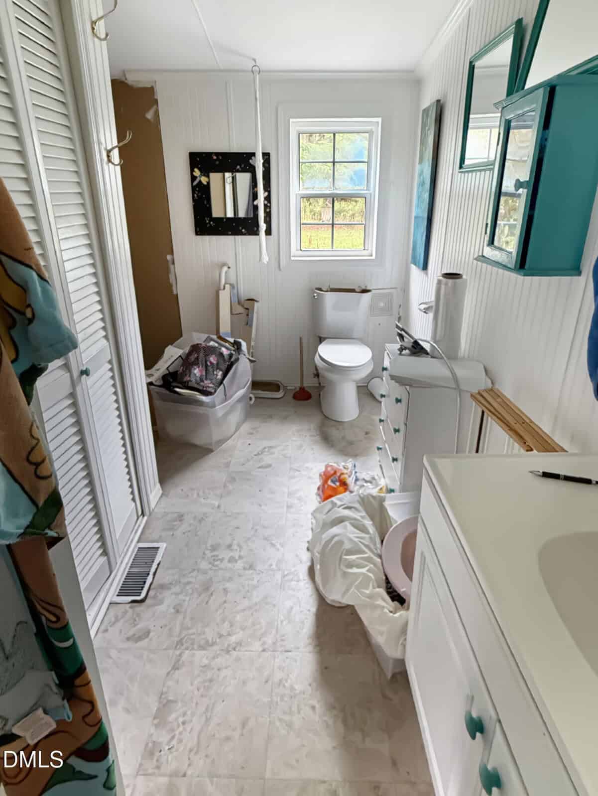 A cluttered bathroom with a toilet, sink, and drawers. Clothes and bags are scattered on the floor. A window lets in natural light. Mirrors and wall art are on the walls. The floor is white tile.