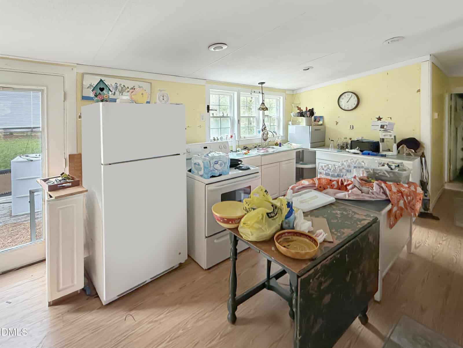 A cluttered kitchen with yellow walls, white appliances, groceries, and dishes scattered on counters and a table. Sunlight comes through windows above the sink.