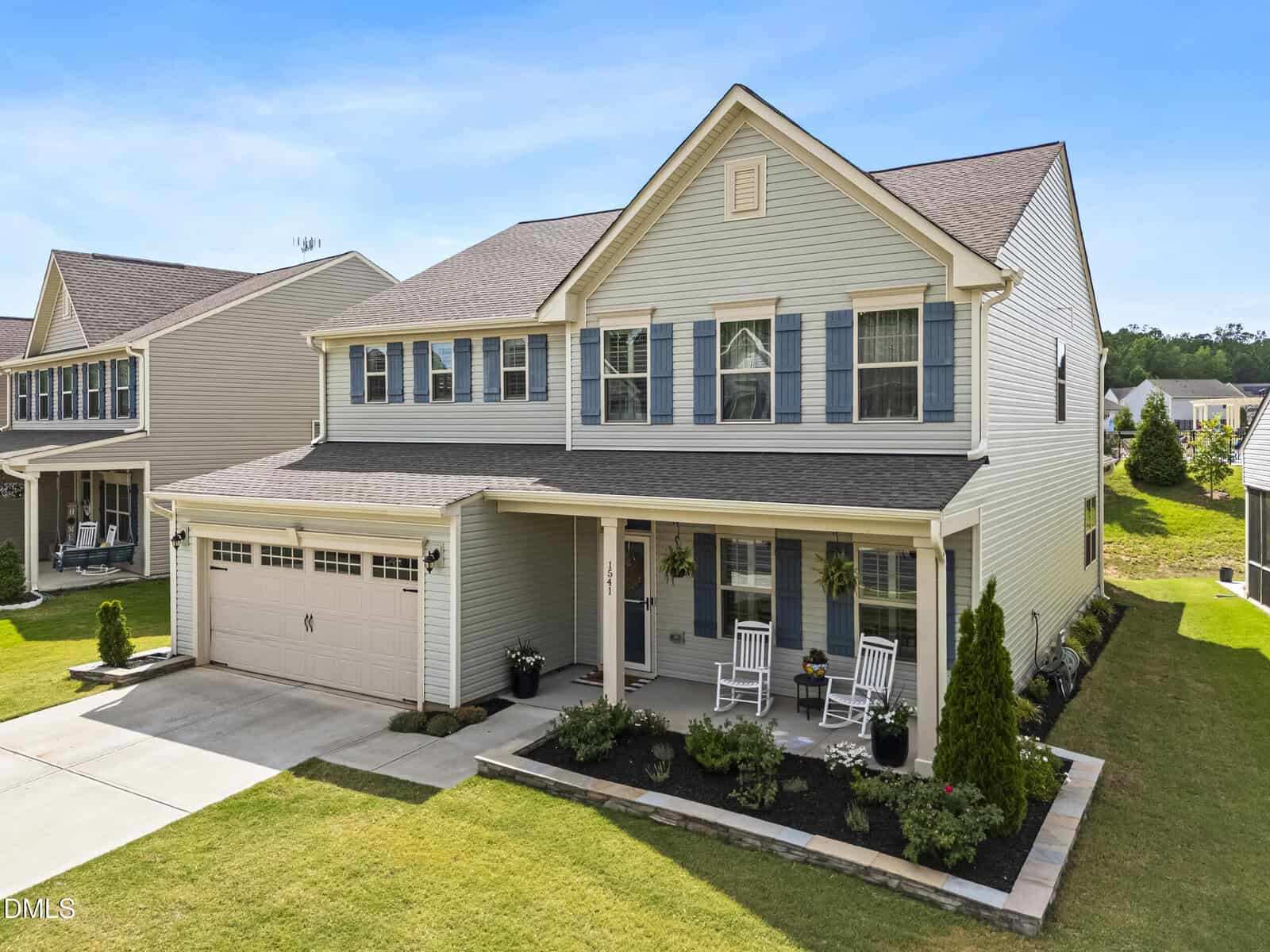 Two-story beige house with blue shutters, a covered front porch, rocking chairs, attached two-car garage, and a well-kept lawn with landscaping, on a sunny day in a suburban neighborhood.