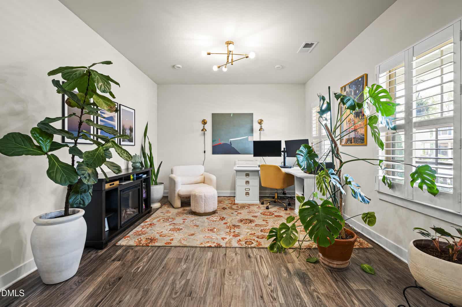 A modern home office with wood floors, indoor plants, a white desk with dual monitors, a white armchair, a decorative rug, wall art, and large windows with shutters letting in natural light.