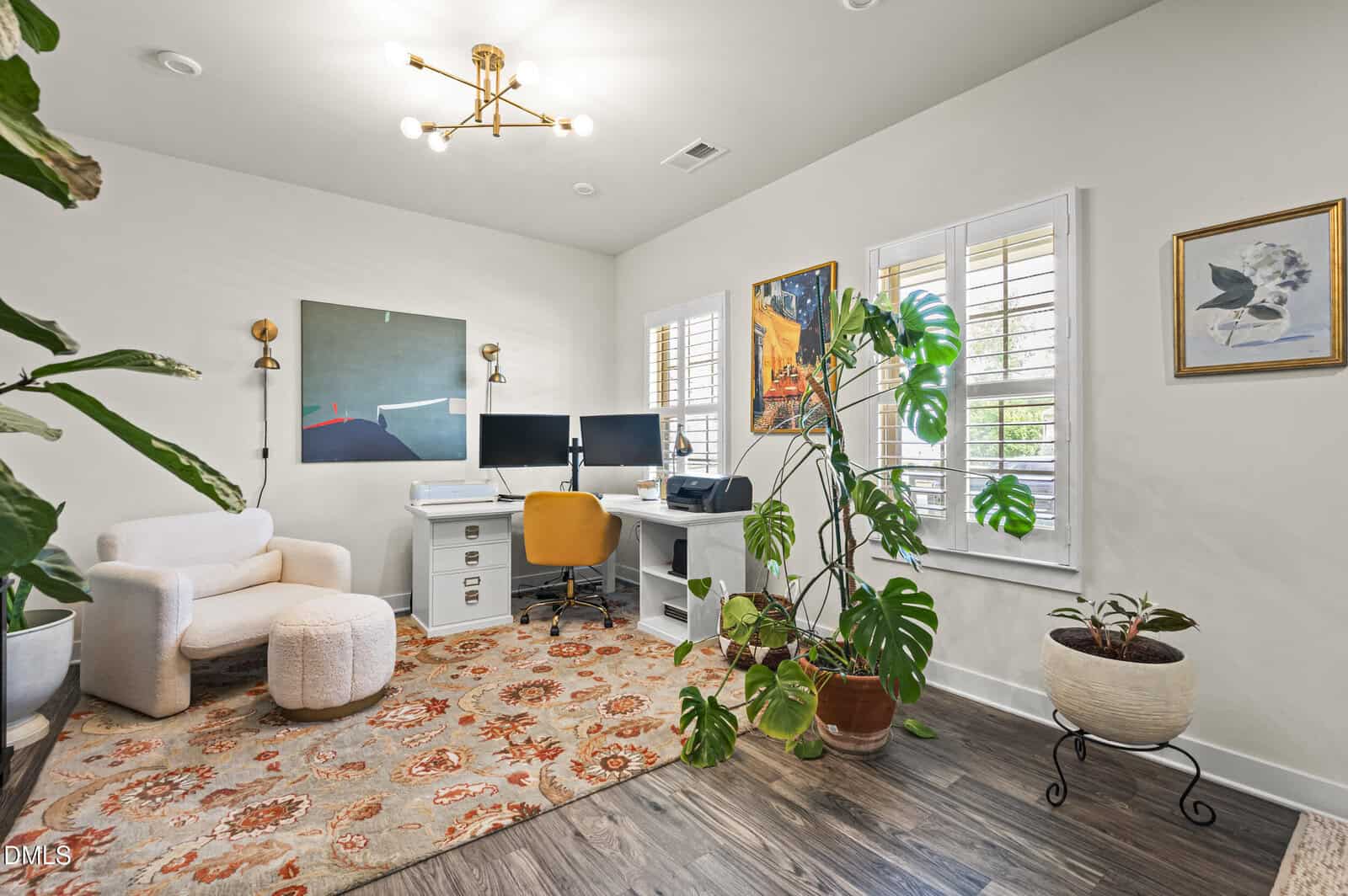 A modern home office with a white desk, computer, yellow chair, and art on the walls. A cozy armchair and patterned rug are in the corner, with large potted plants near shuttered windows letting in natural light.
