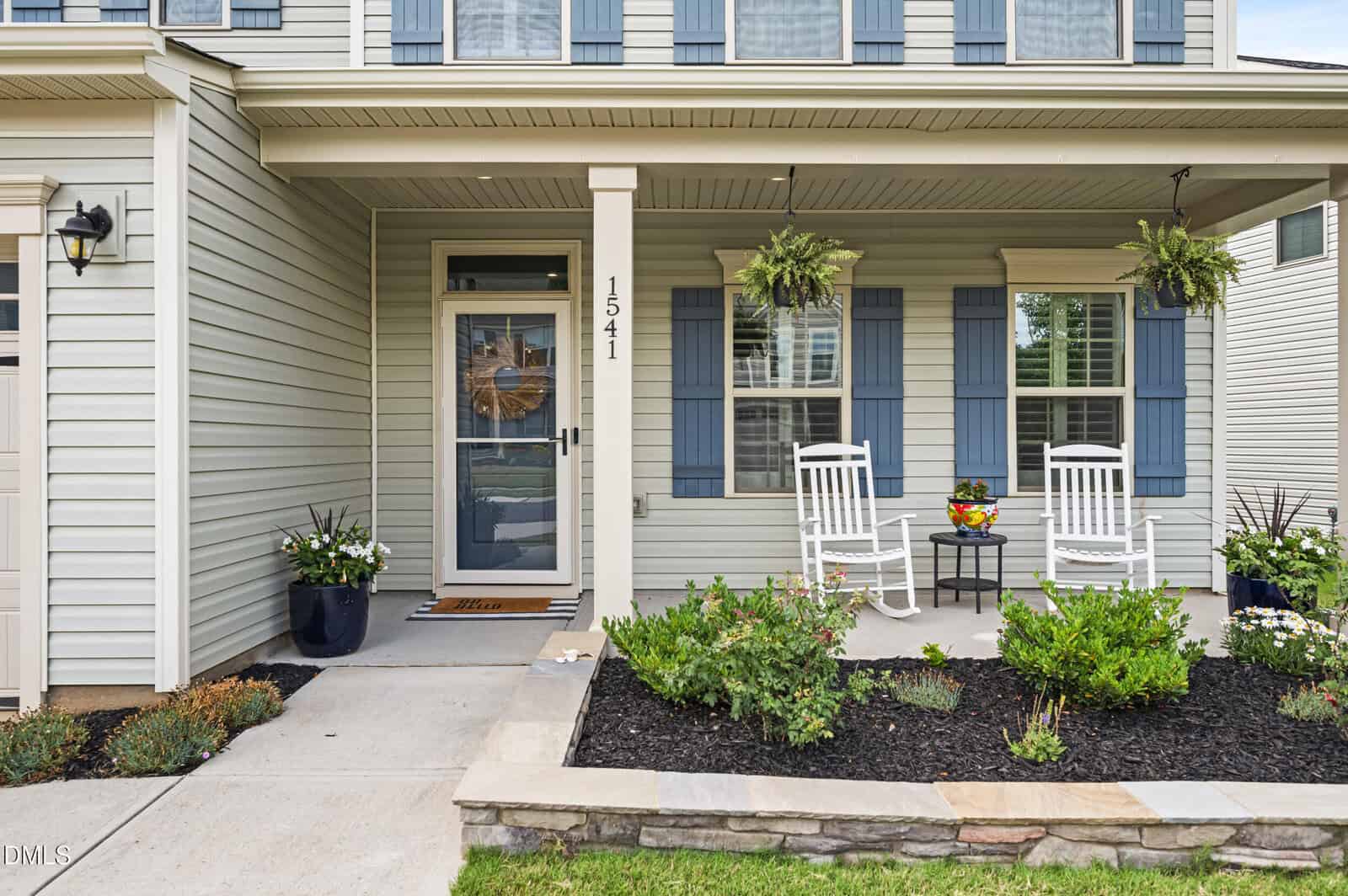 Front porch of a light-colored house with blue shutters, a glass front door, two white rocking chairs, potted plants, hanging ferns, and landscaped flower beds along a stone-bordered walkway.