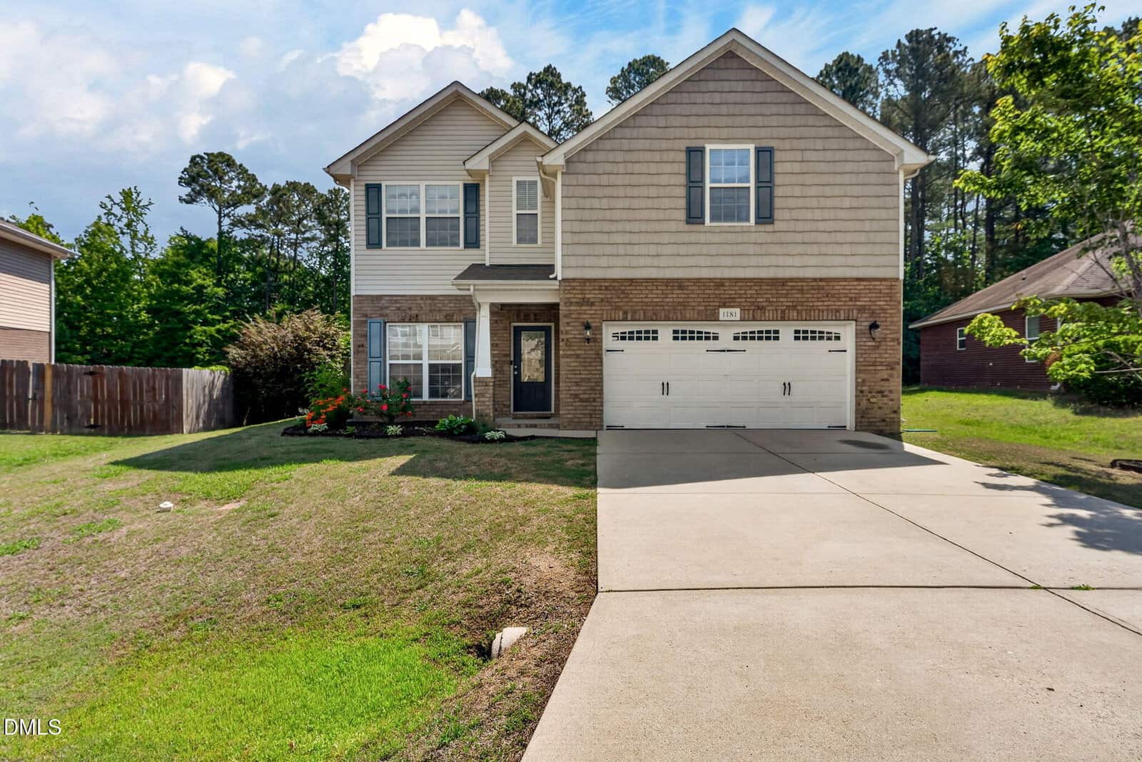 Two-story suburban house with beige siding, brick accents, and a double garage. The front yard has green grass, shrubs, and a small flower garden. Tall trees and neighboring houses are in the background under a partly cloudy sky.