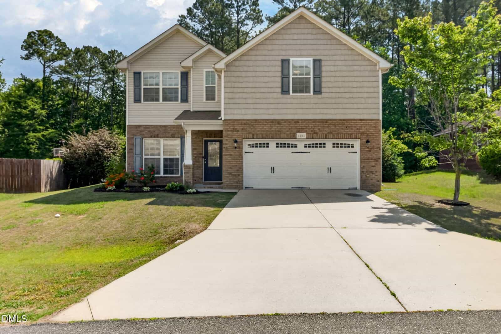 Two-story suburban house at 1181 Ridgeland Drive in Creedmoor, featuring beige siding, a brick facade, and a white double garage. A driveway leads to the entrance, with a lawn bordered by plants and trees under a partly cloudy sky.