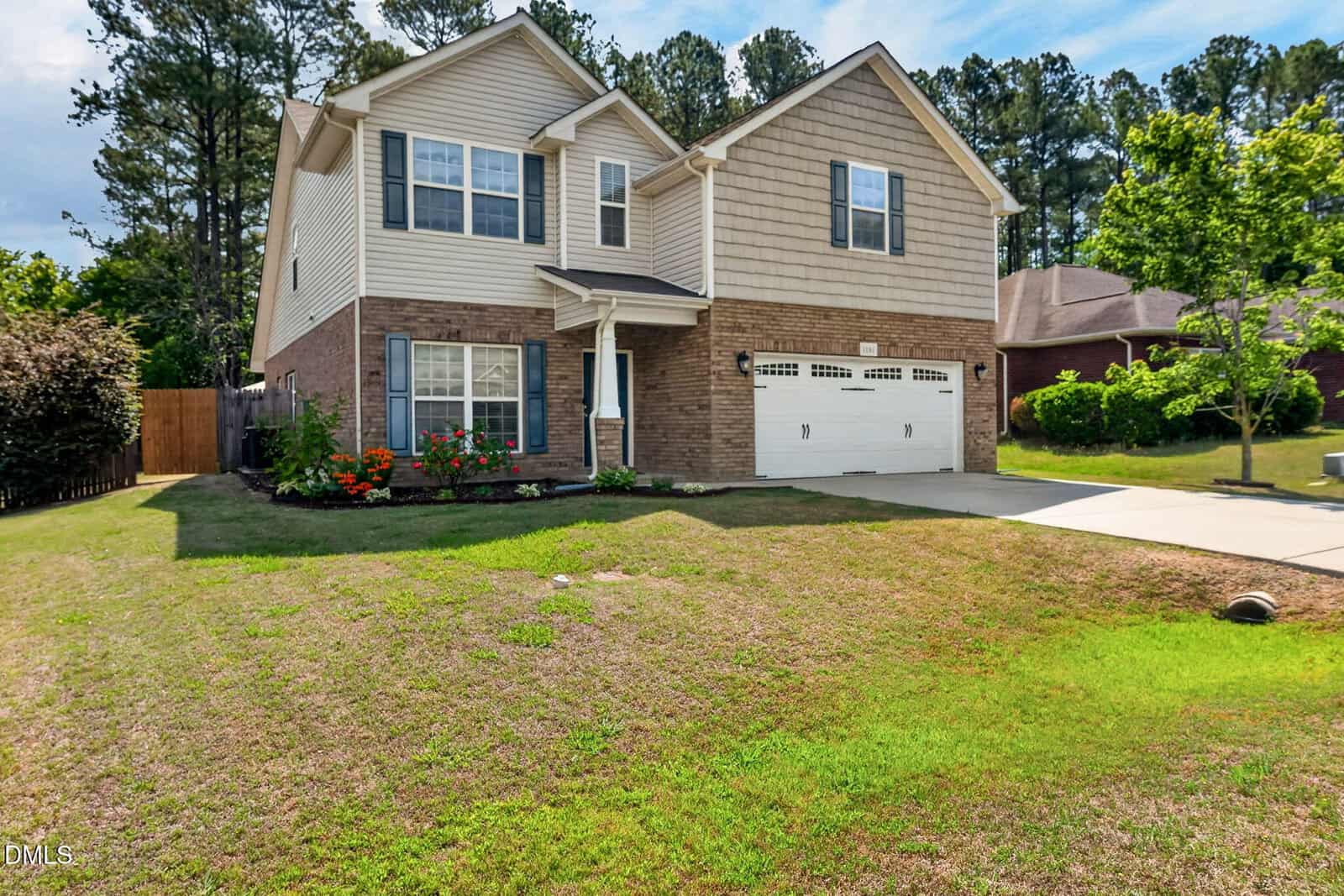 Two-story suburban house with brick and beige siding, attached two-car garage, flower beds near the entrance, and a driveway. The front lawn has patches of green and brown grass, with trees and shrubs in the background.