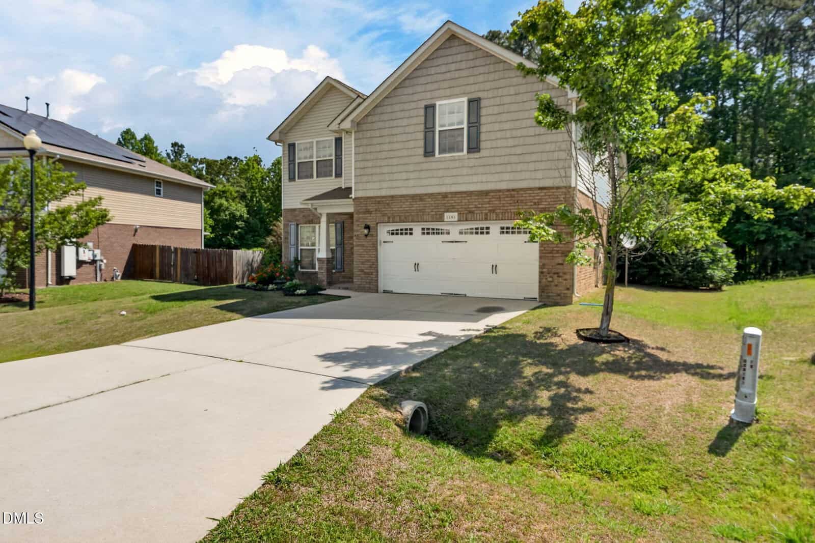 A two-story suburban house with a double garage, light brown brick and siding exterior, a concrete driveway, and a grassy yard bordered by trees under a partly cloudy sky.