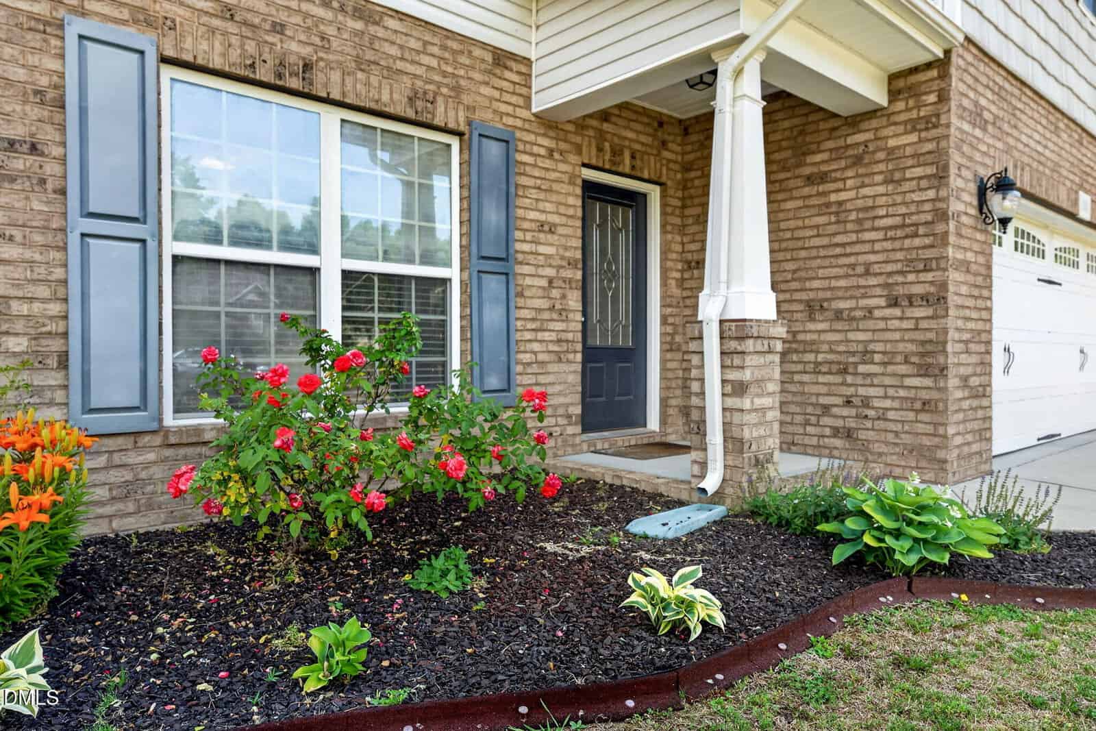 A brick house exterior features a black front door, large windows with white trim, a small porch with a white column, and a well-maintained flower bed with blooming red and orange flowers and green plants.