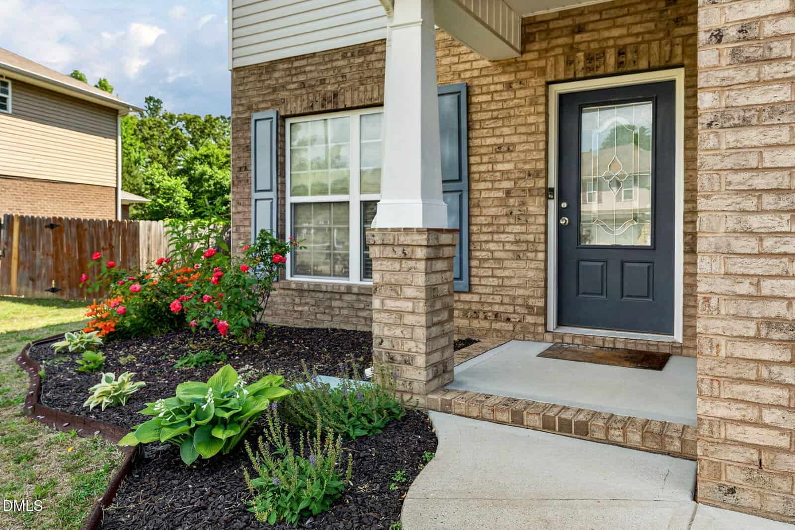 A brick house entryway with a black door, glass panel, and a small porch. The walkway curves past a flower bed with red and green plants, bordered by mulch. A wooden fence and neighboring house are in the background.