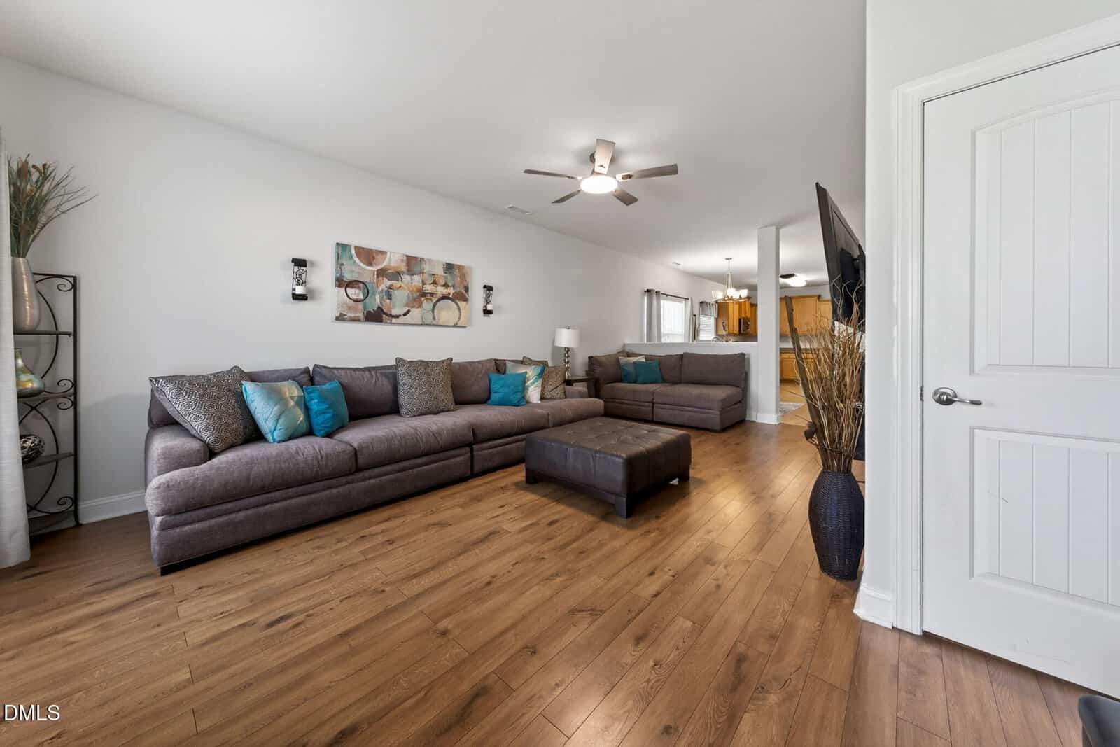 Modern living room with hardwood floors, gray sectional sofa, blue and brown accents, ottoman, wall art, ceiling fans, and a view into the kitchen at the back. White walls and a closed white door are visible.