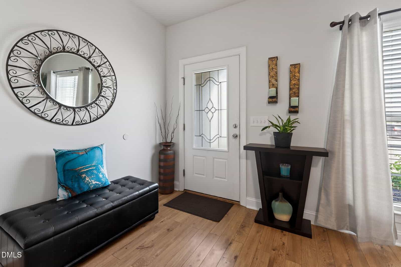 Entryway with a white door, circular mirror, black bench with a blue pillow, tall vase, dark console table with decor, and white curtains over a window. Light wood floor throughout.