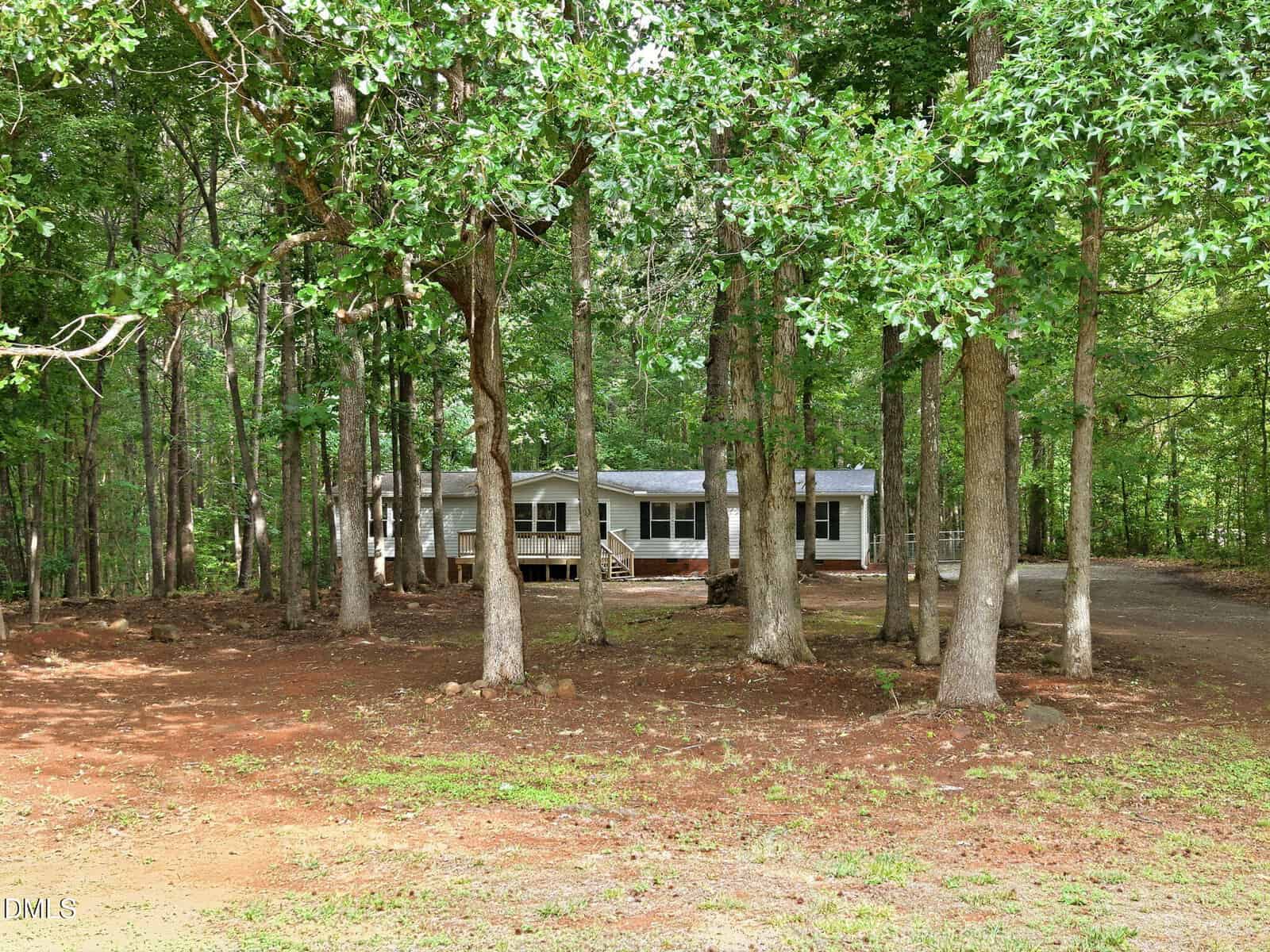 A white, single-story house with a porch sits in a wooded area at 3593 Crosswinds Drive, partially obscured by tall trees. The ground is mostly bare with patches of grass, and a dirt driveway curves toward the house.