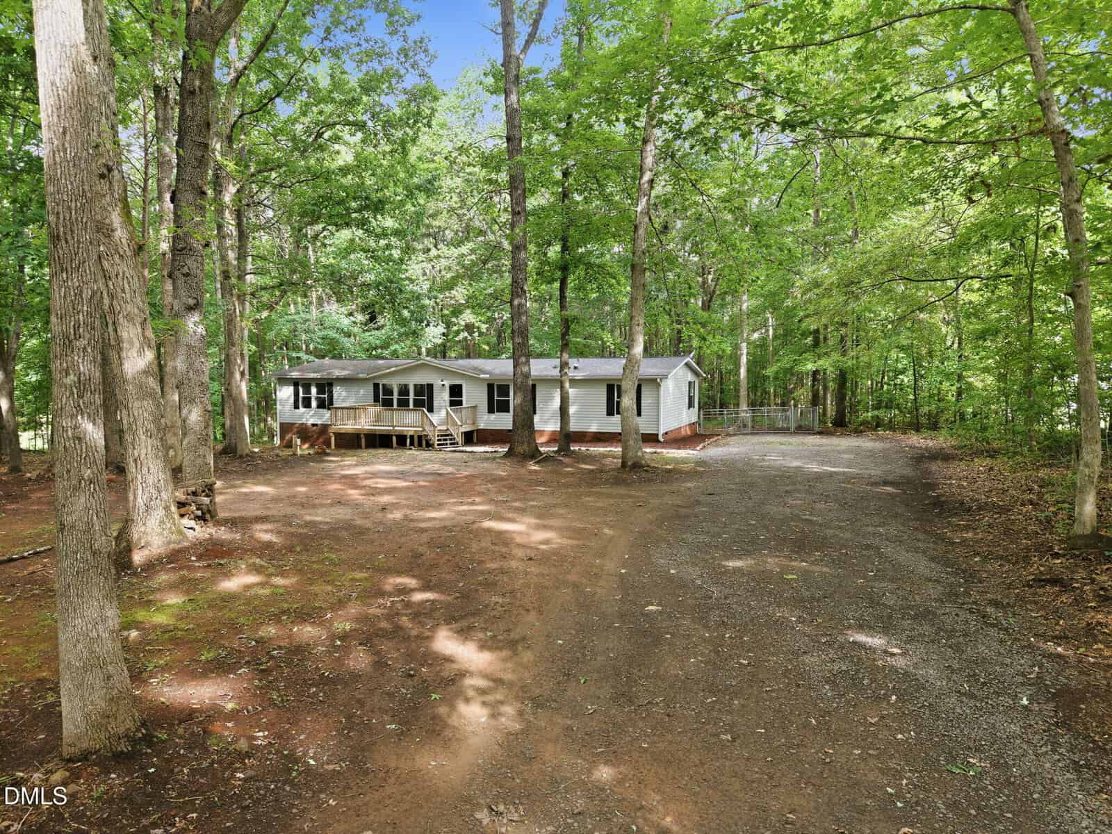 A white single-story house with a green roof is surrounded by tall trees, with a dirt and gravel driveway curving in front, set in a wooded area on a sunny day.