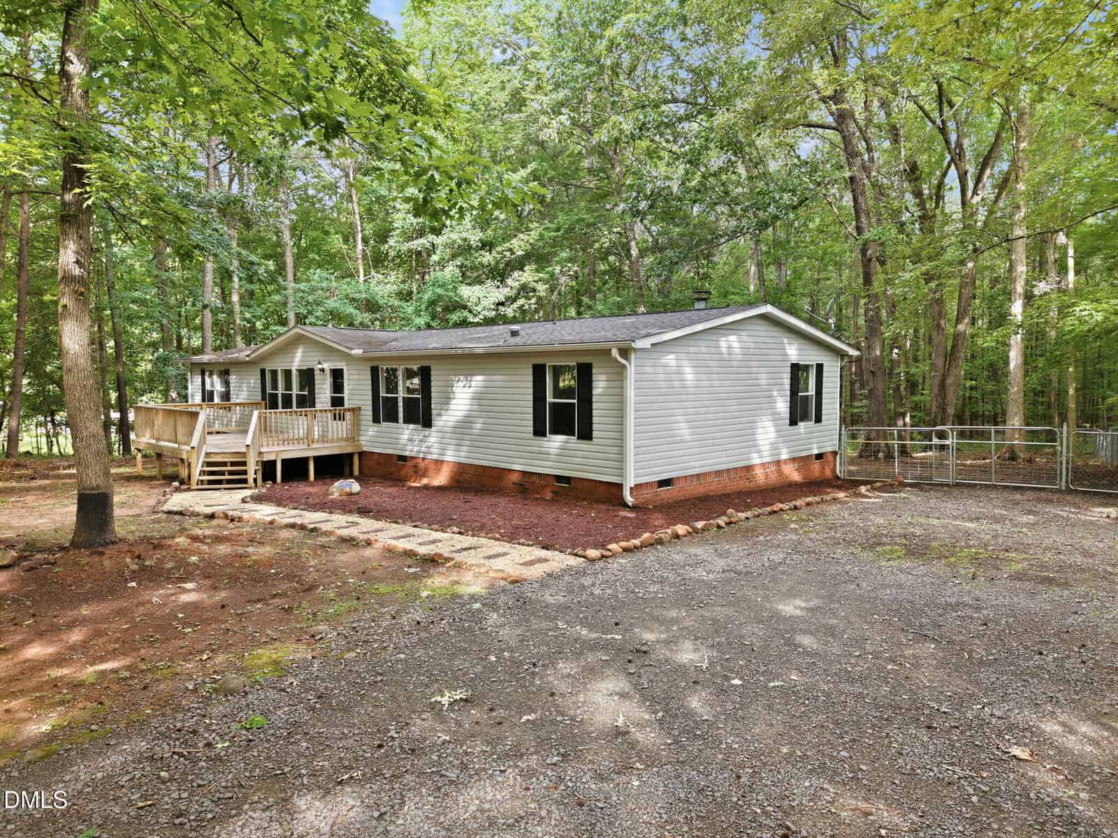 A single-story white house with black shutters sits among tall trees. It has a small wooden porch, a brick foundation, and a gravel driveway. The yard is partially mulched with a walkway leading to the entrance.