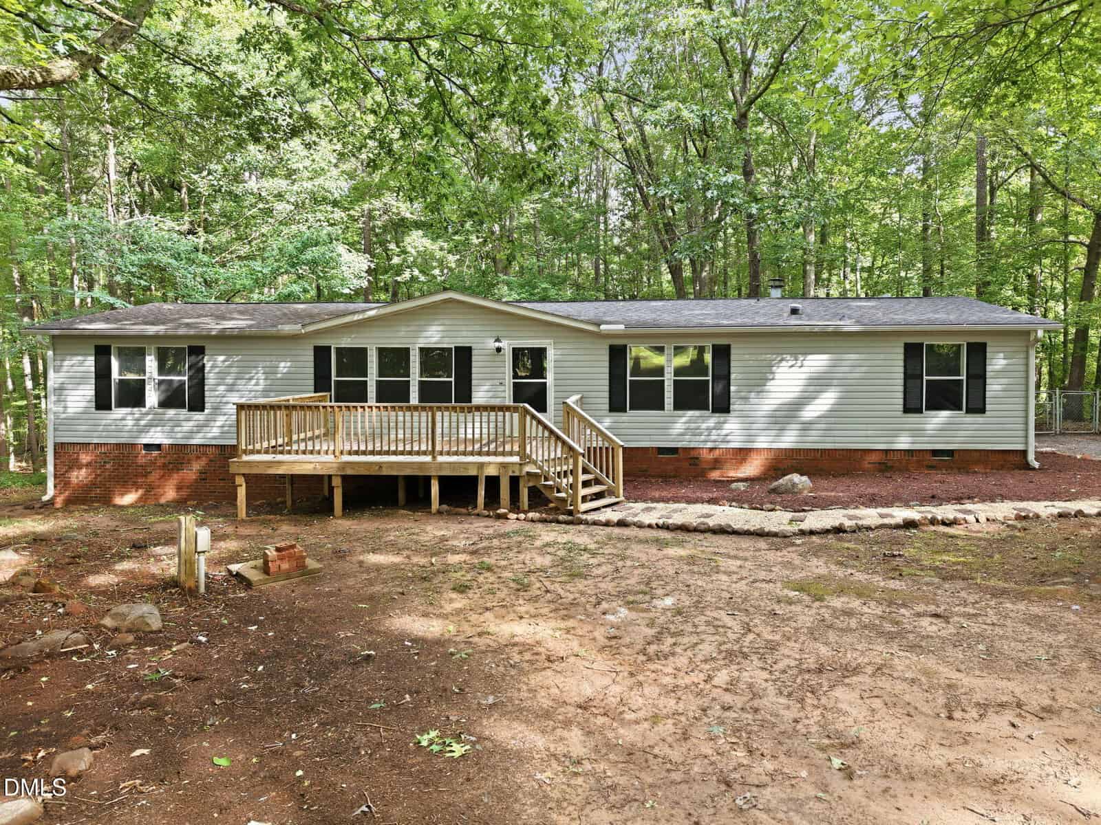 Single-story house with light siding, dark-framed windows, and a brick foundation, surrounded by trees. A wooden ramp and steps lead to the front door, and the yard has a dirt surface with scattered rocks.