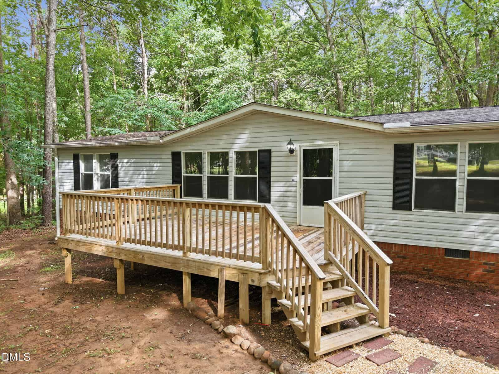 A single-story house with light gray siding, black shutters, and a wooden ramp-style front porch surrounded by trees and a grassy yard. The porch has railings and steps leading to the front door.