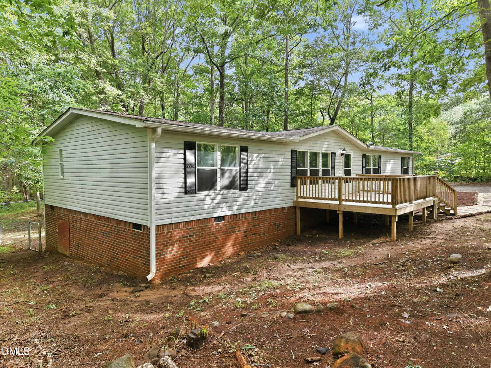 Single-story house with white siding and a brick foundation, featuring black shuttered windows and a wooden deck, surrounded by trees and natural dirt ground.
