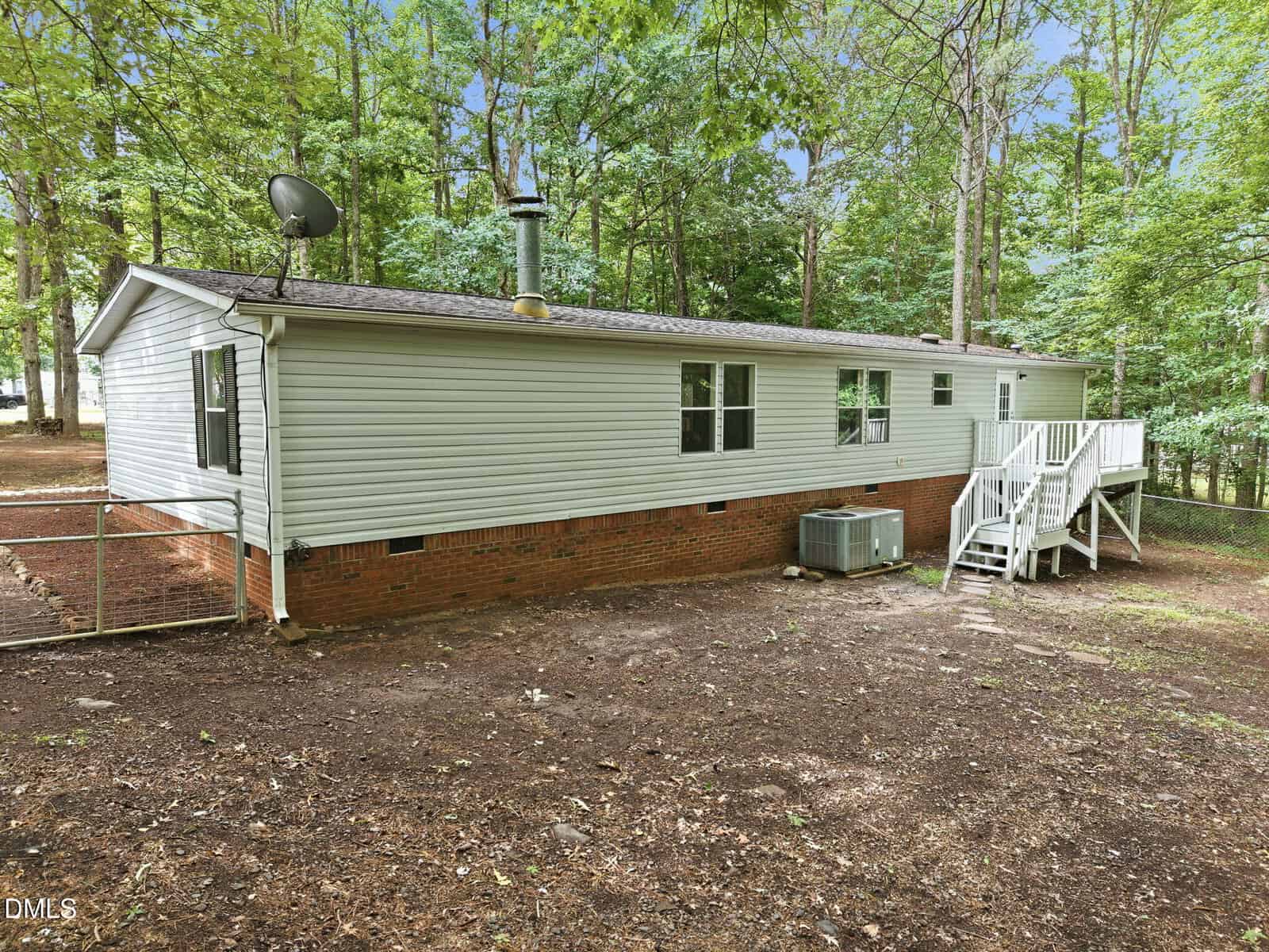 Single-story house with light-colored siding, brick foundation, and rear steps leading to a white door, surrounded by trees and bare ground in the backyard. Satellite dish and AC unit are visible outside.