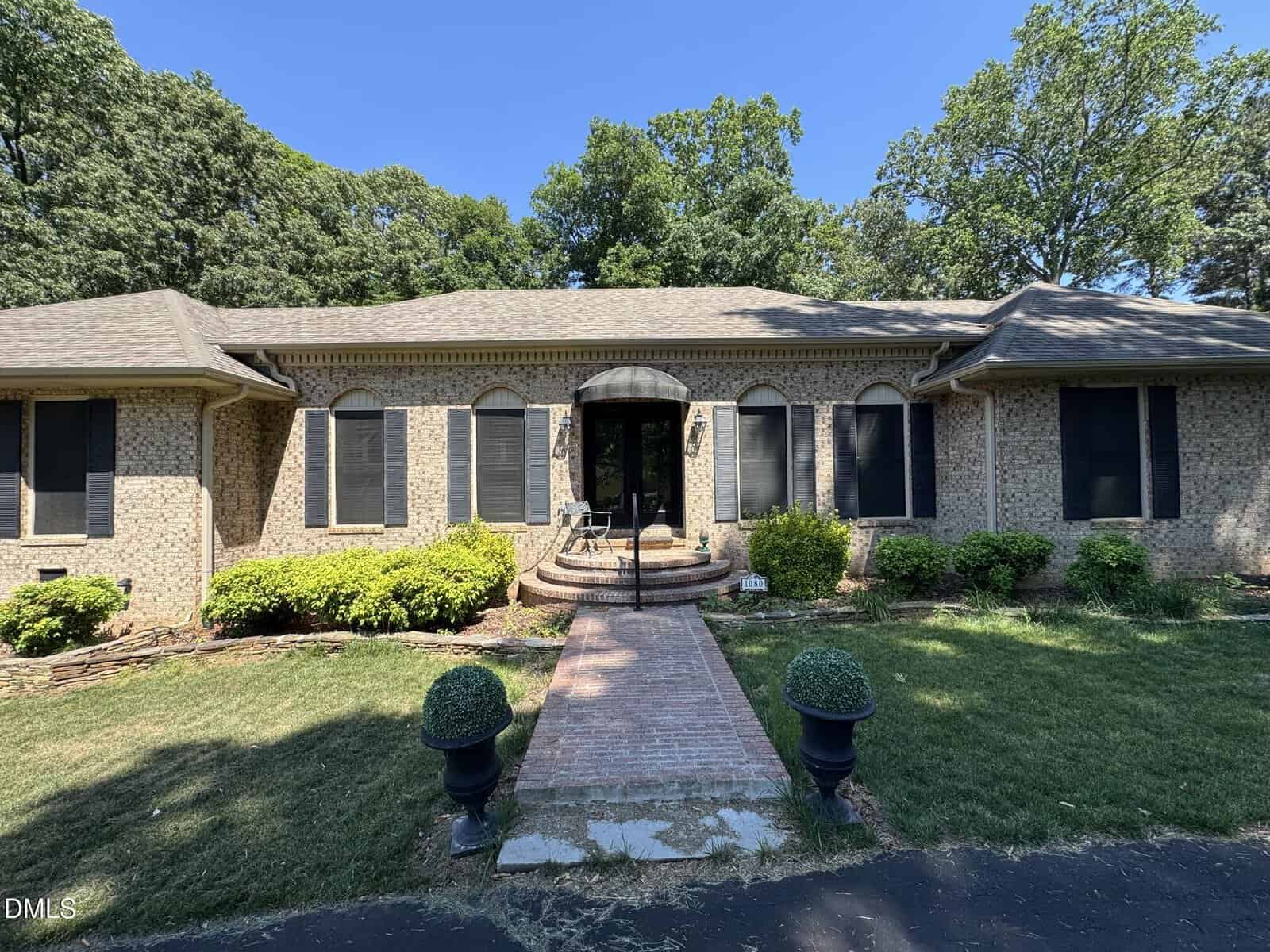 Single-story brick house with black shutters and a central arched doorway. A brick walkway leads to the entrance, flanked by trimmed bushes and two potted topiary plants. The house is surrounded by green lawn and trees.