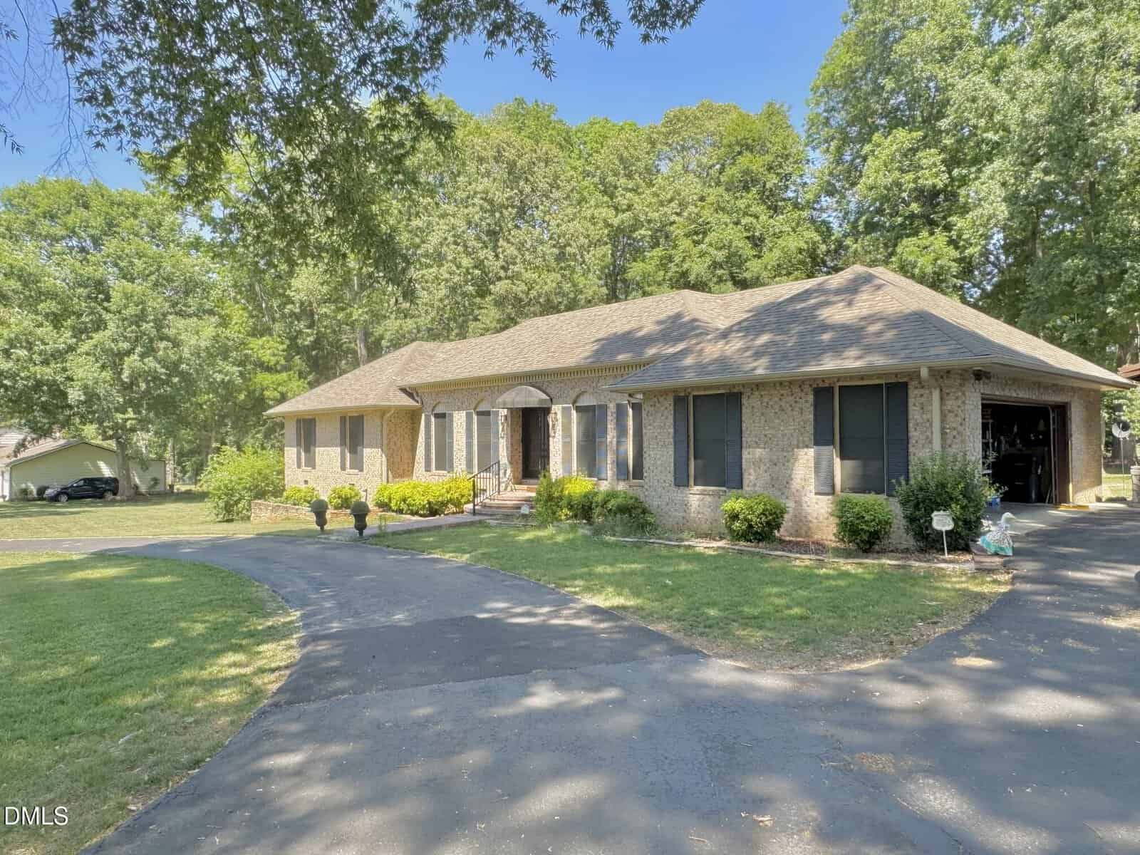 Single-story brick house with a gabled roof, black shutters, and a curved driveway. The yard is well-kept with green grass, shrubs, and trees in the background under a clear blue sky.