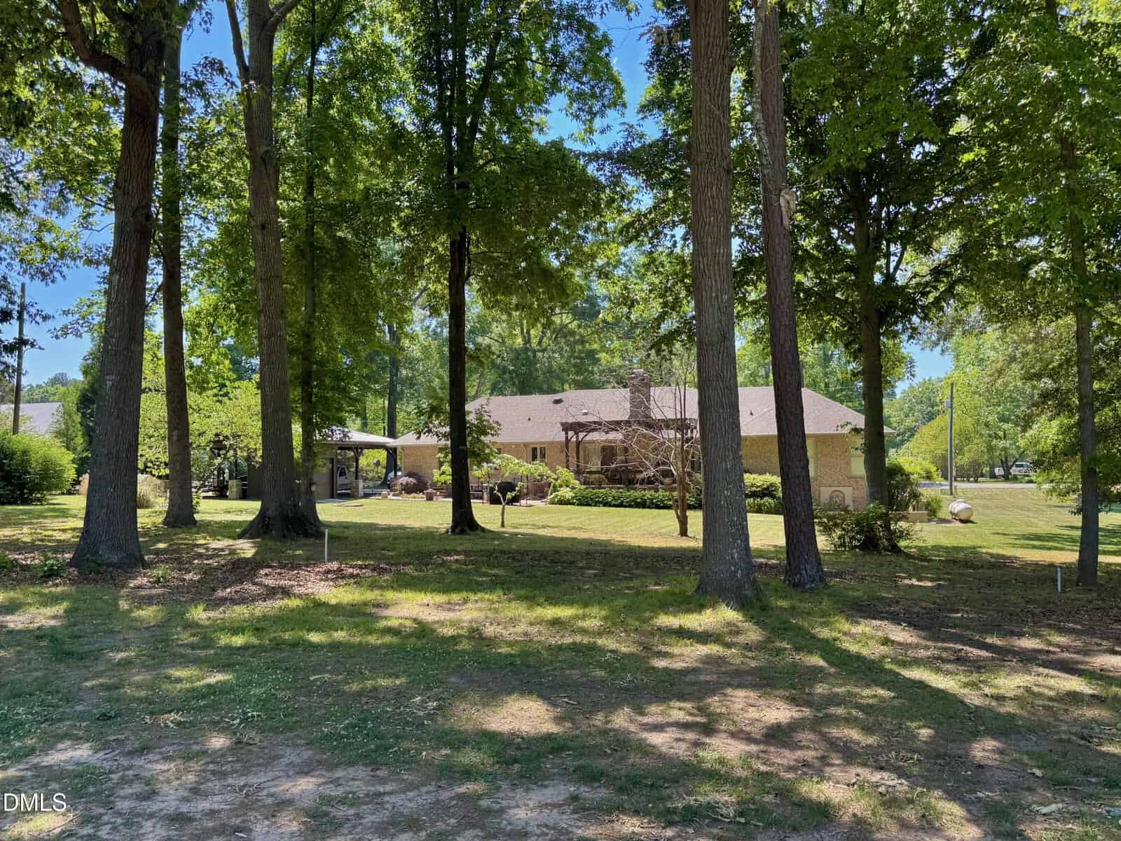 A single-story house with a beige brick exterior is partly visible through tall trees on a sunny day, surrounded by green grass, shrubs, and a shaded yard.