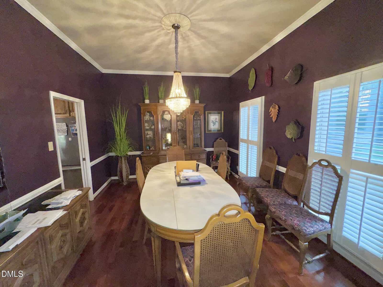 A dining room with a long oval table, ten chairs, a crystal chandelier, dark purple walls, large plants, wall art, a china cabinet, and windows with white shutters. Papers and binders are on the table.