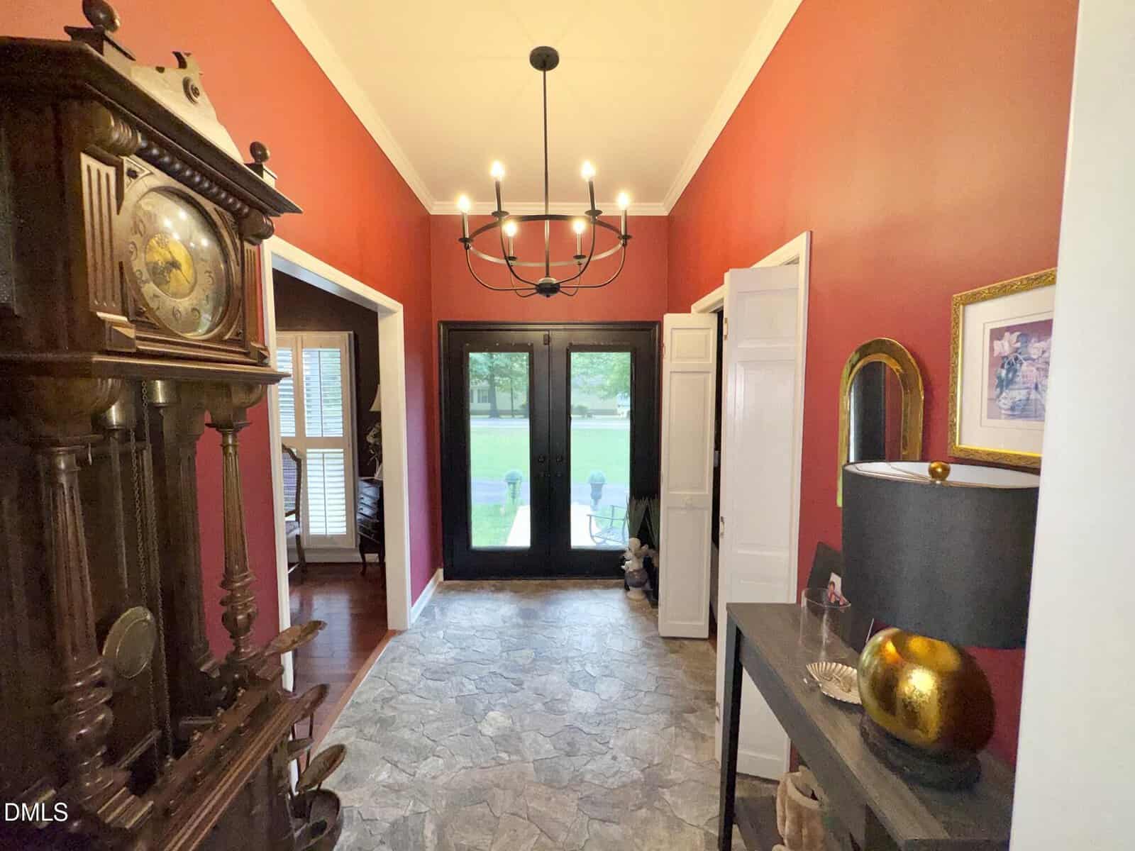A hallway with red walls, a stone tile floor, a black chandelier, a grandfather clock, double front doors with glass panels, a console table with a black lamp, and framed art. Patio chairs are visible outside the doors.