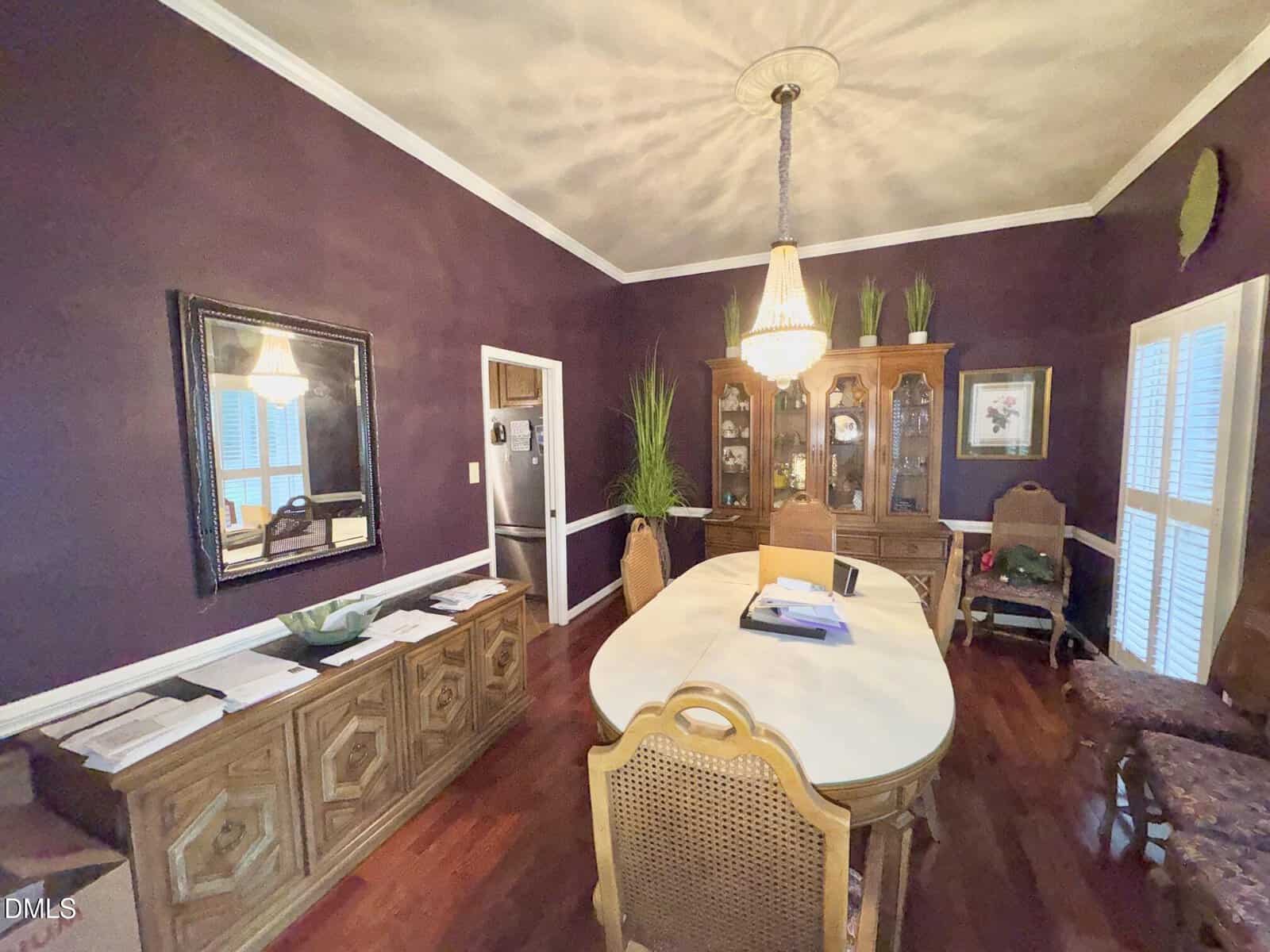 A formal dining room with dark purple walls, wooden furniture, a long oval table, chandelier, hutch with plants, and papers on the sideboard. Light enters through tall windows with white shutters.