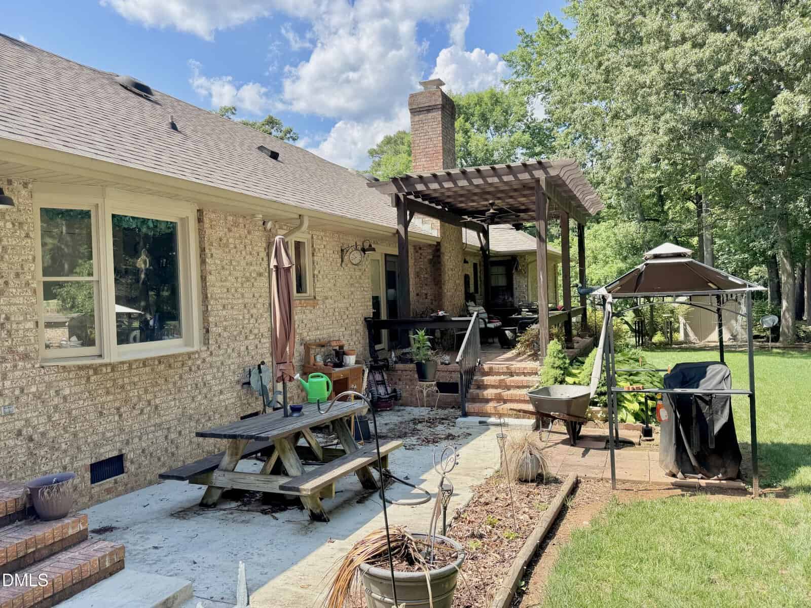 A backyard with a brick house, covered patio with pergola, picnic table, garden tools, wheelbarrow, and gazebo, surrounded by green grass and trees under a partly cloudy sky.