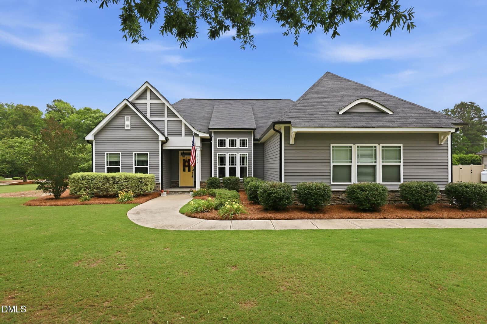 A modern gray house with white trim at 15 Ashland Court, Zebulon, features a gabled roof and large windows behind manicured bushes and a curving sidewalk, surrounded by green lawn and trees under a blue sky.