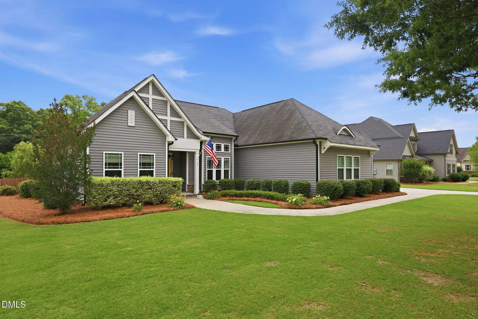 A modern single-story house with gray siding, white trim, and a gabled roof stands on a well-manicured lawn with shrubs, mulch beds, and an American flag by the front entrance under a blue sky.
