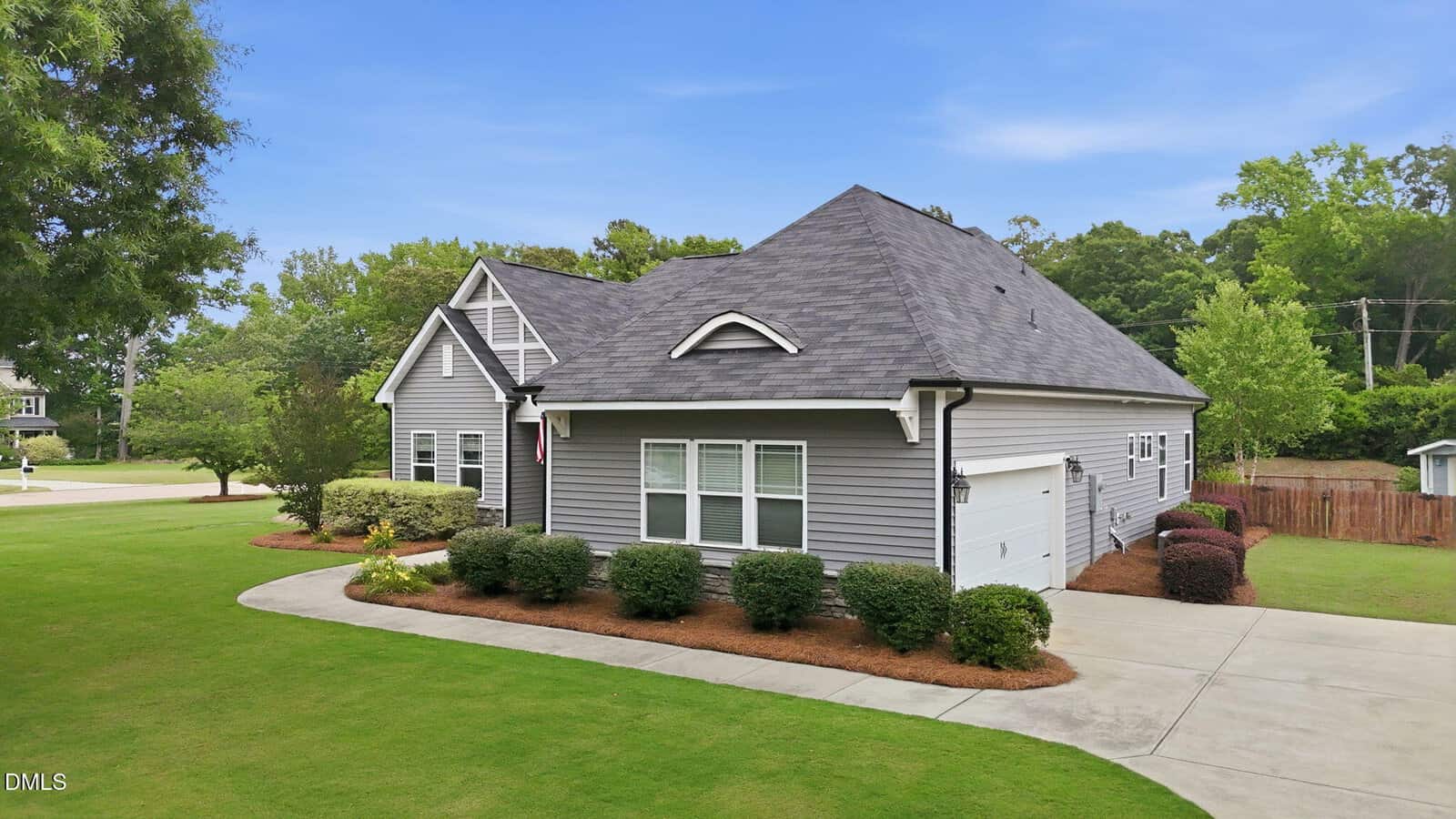 A gray single-story house with white trim, a two-car garage, and manicured landscaping stands on a corner lot with green grass, shrubs, and trees under a blue sky.