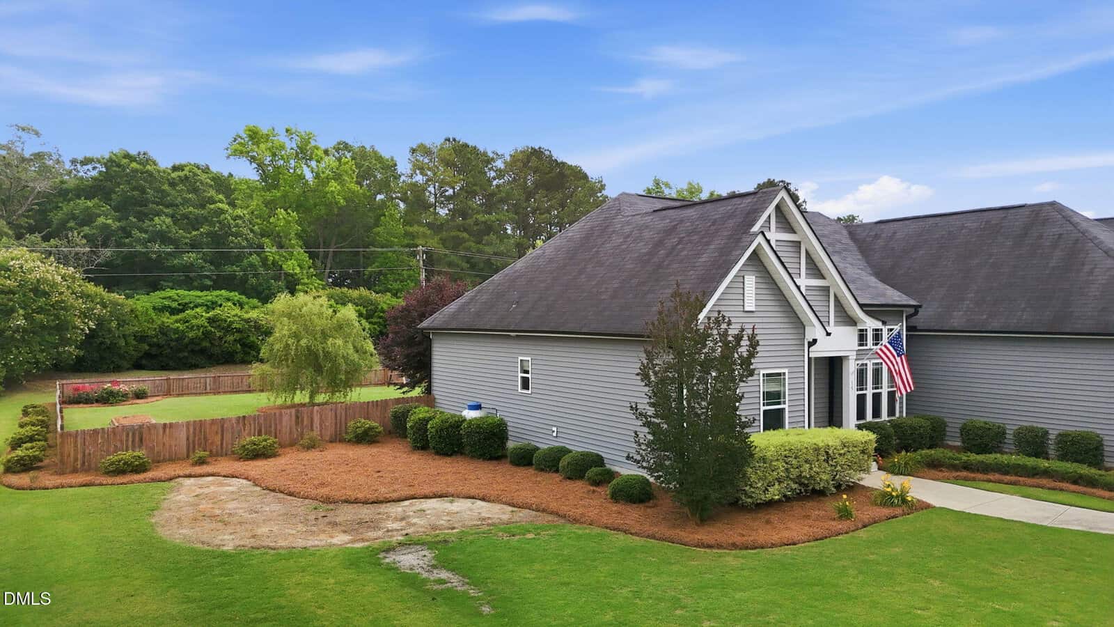 A gray house with white trim sits on a well-maintained lawn, featuring neatly trimmed bushes, a fenced backyard, and an American flag by the entrance. Blue sky and green trees are visible in the background.