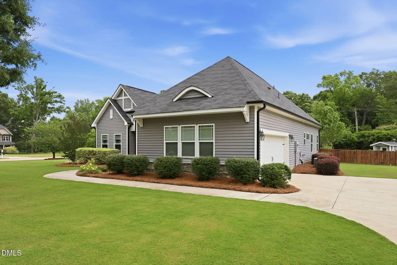 A single-story gray house with white trim, a large front lawn, landscaped shrubs, and a concrete driveway under a blue sky with scattered clouds.