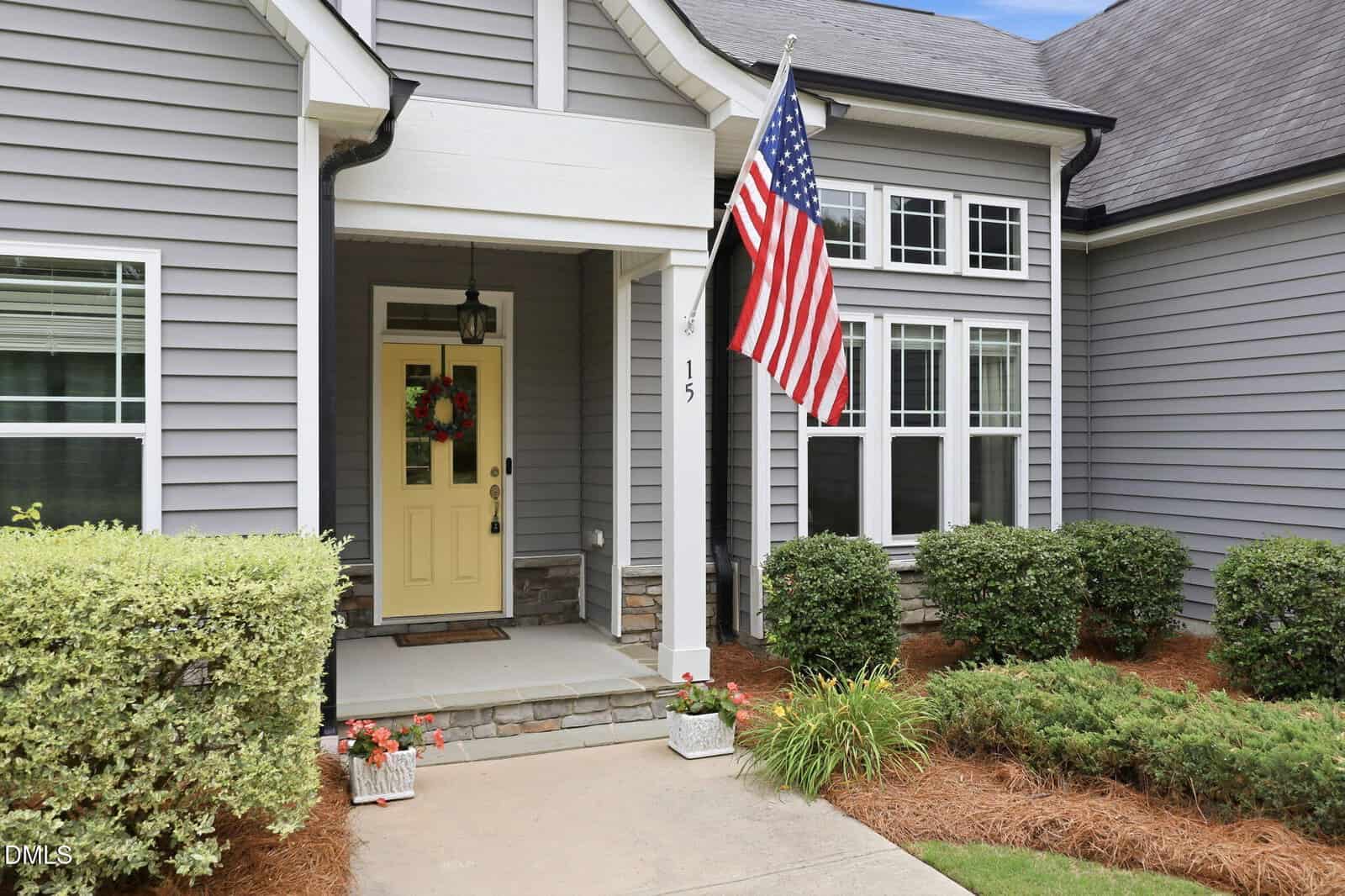 Gray house with a yellow front door, American flag, and wreath. Well-kept bushes and mulch line the walkway. Steps lead to the covered porch with potted flowers by the entrance.