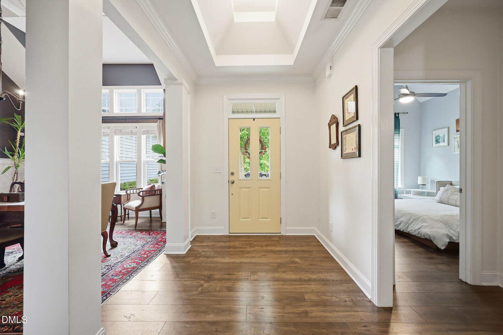 Bright entryway with yellow door, wooden floor, and white walls. Hallway leads to a bedroom on the right and a dining room with a red rug and large windows on the left. Framed art hangs on the hallway wall.
