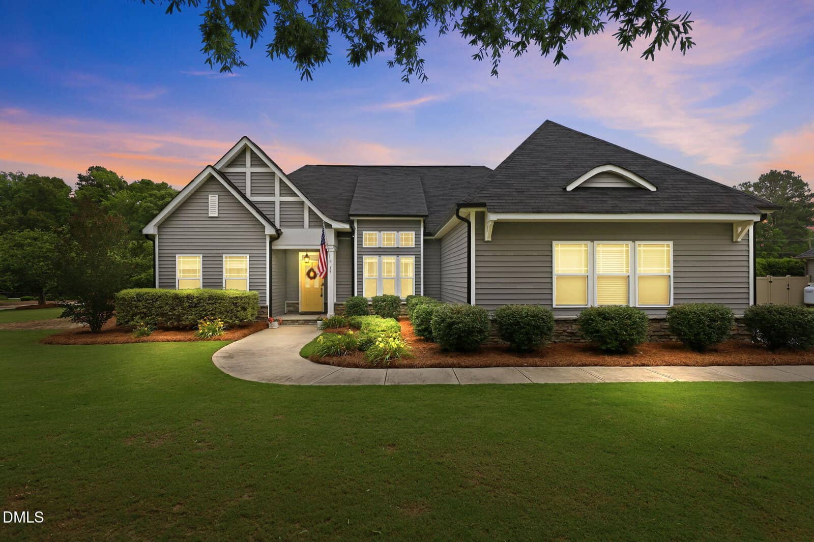 A single-story gray house with white trim, neatly manicured lawn, shrubs, and outdoor lights glowing at dusk. An American flag hangs near the front door, and a curved walkway leads to the entrance.