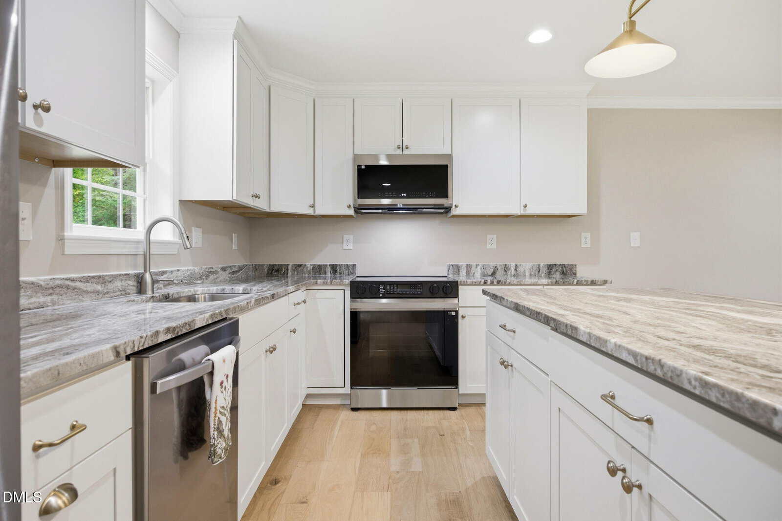 Modern kitchen with white cabinets, marble countertops, stainless steel appliances, a built-in oven, microwave, and light wood floors. A window over the sink lets in natural light.