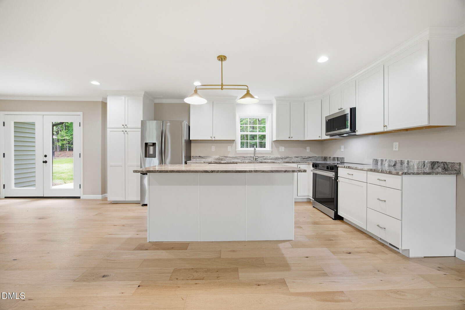 Modern kitchen with white cabinets, marble countertops, stainless steel appliances, a central island, light wood floors, pendant lighting, and double doors leading to a green backyard.