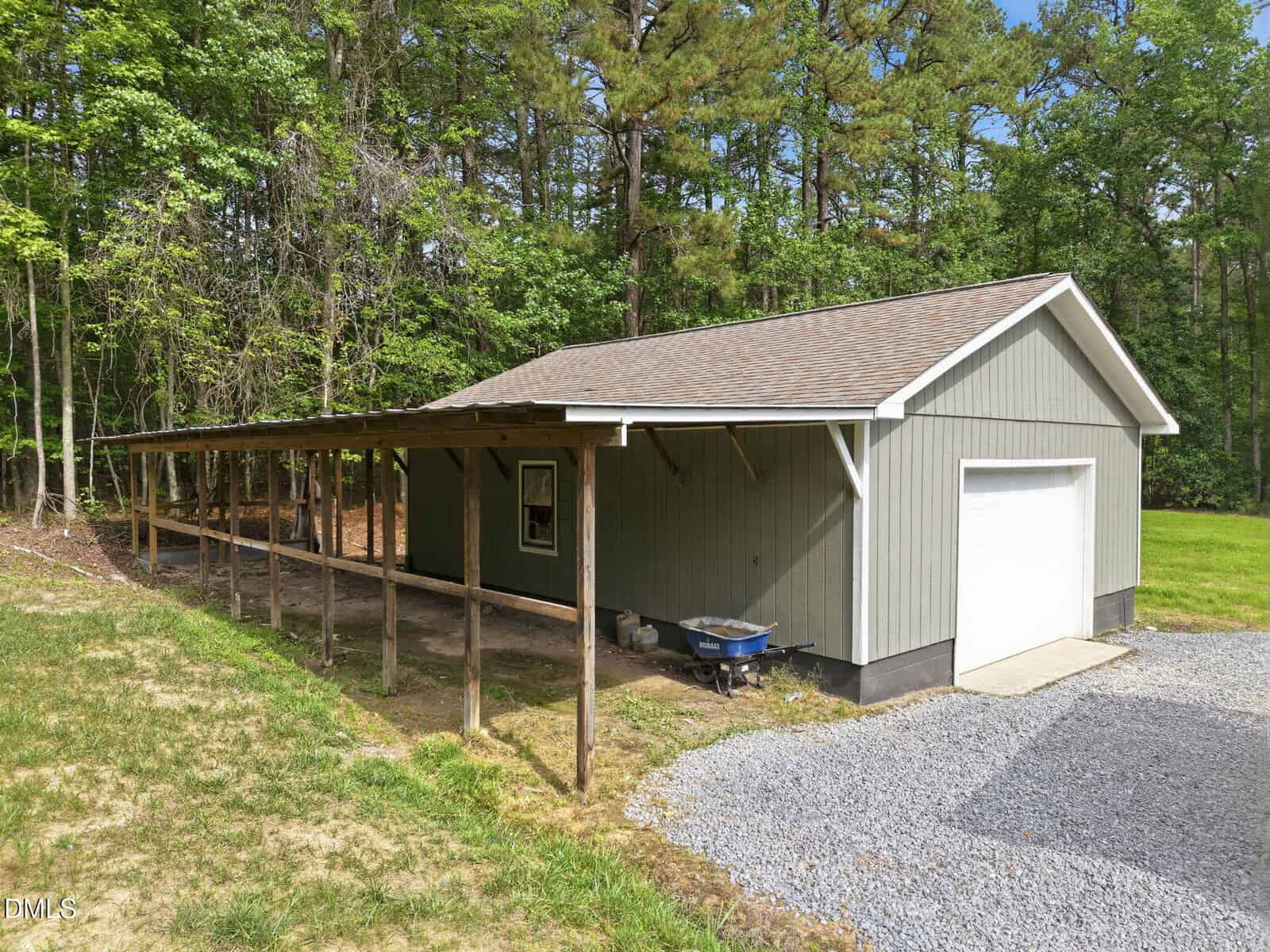 A gray wooden shed with a gable roof, white trim, and a white garage door sits next to a gravel driveway. An attached wooden carport runs along one side. The building is surrounded by trees and grass.