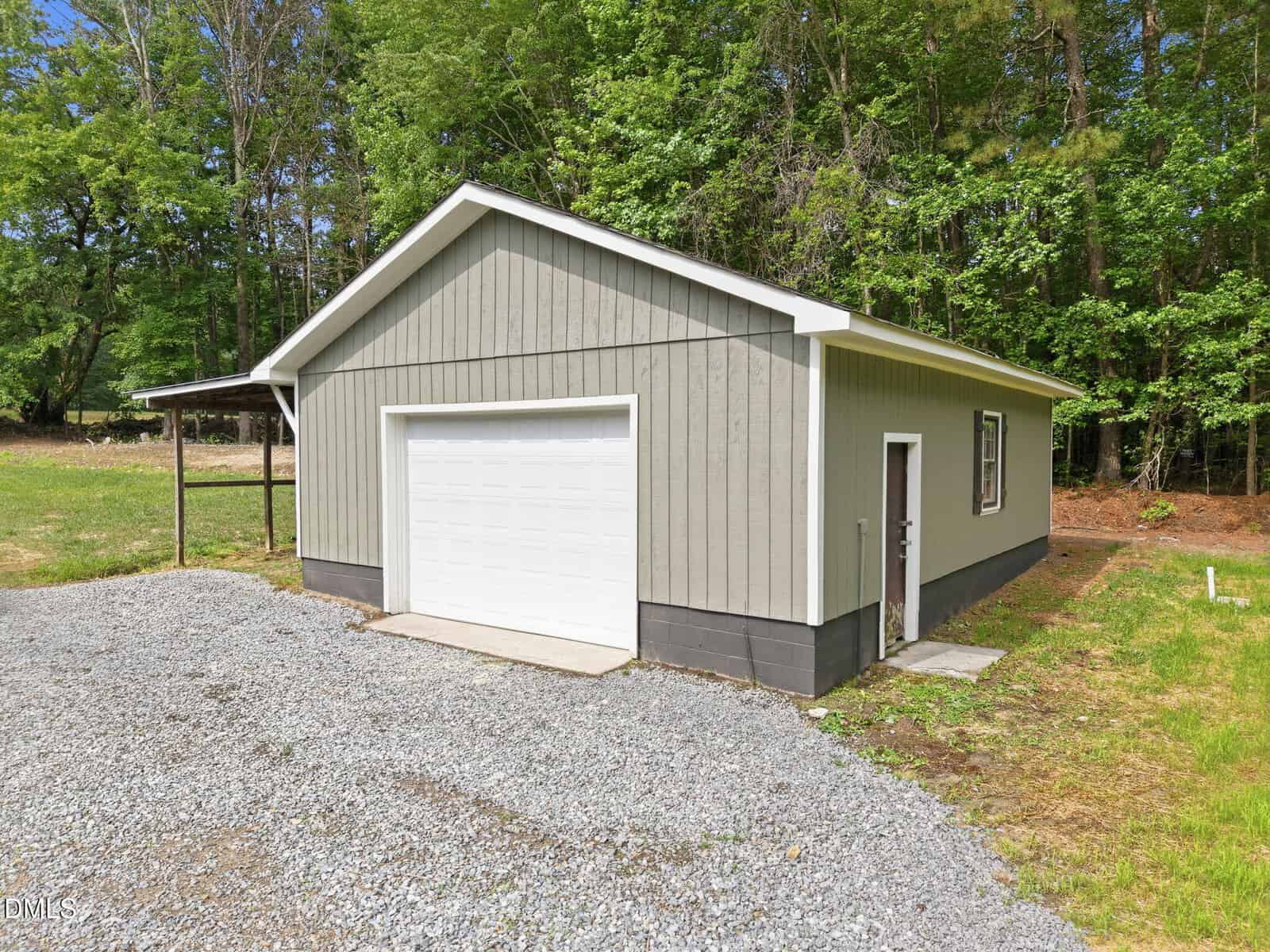 A small, gray detached garage with a white door sits on a gravel driveway surrounded by grass and trees under a clear sky.